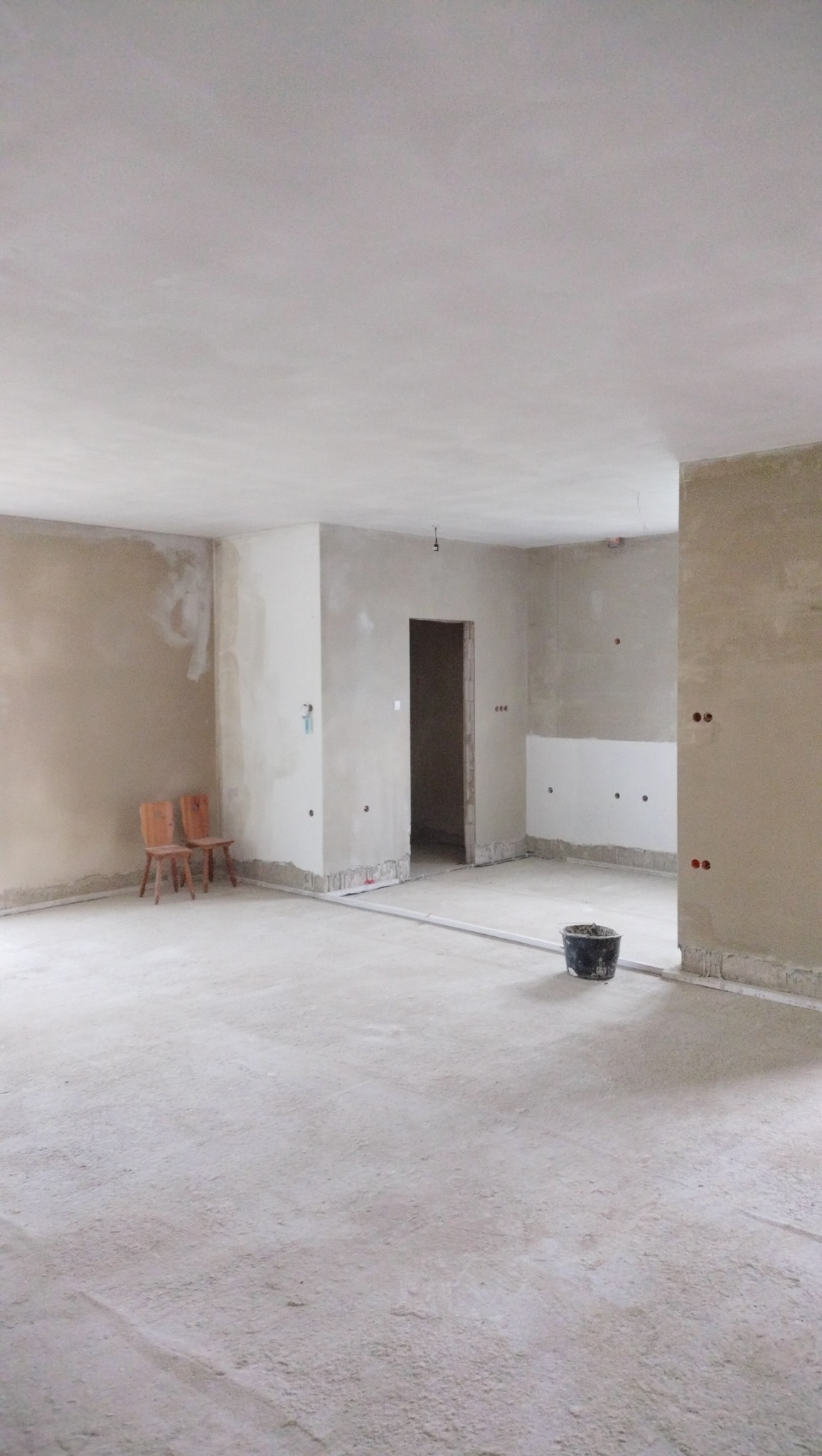 Surowe, jasne wnętrze w trakcie remontu: ściany po wstępnym wyrównaniu, widoczne ślady po szpachlowaniu, plastikowe listwy przypodłogowe, dwa krzesła przy ścianie i wiadro na podłodze.