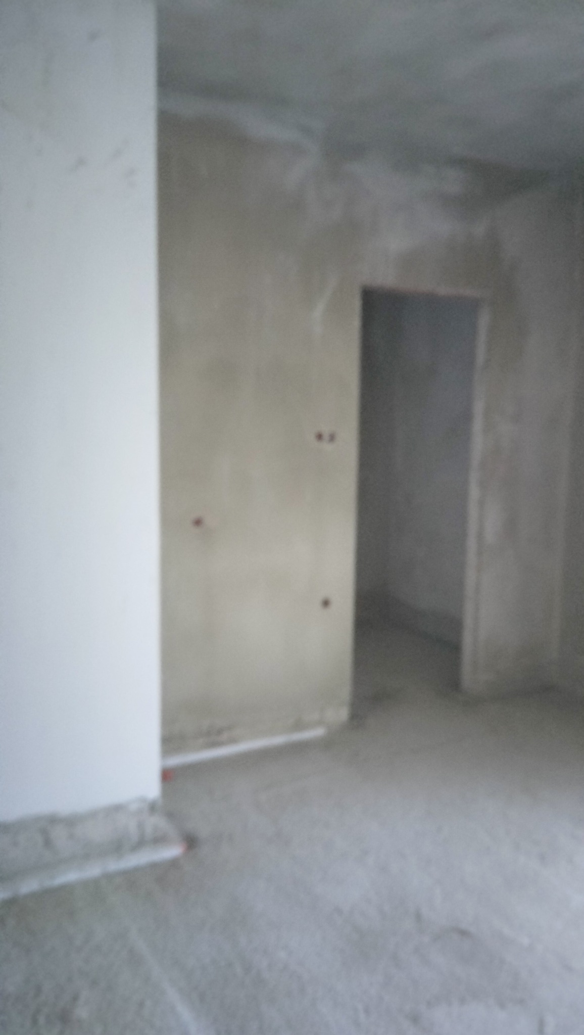 Surowe wnętrze w trakcie prac wykończeniowych: ściany z widocznymi śladami zaprawy, przygotowane do dalszych prac, widoczne otwory po instalacjach elektrycznych, podłoga pokryta warstwą wylewki,...