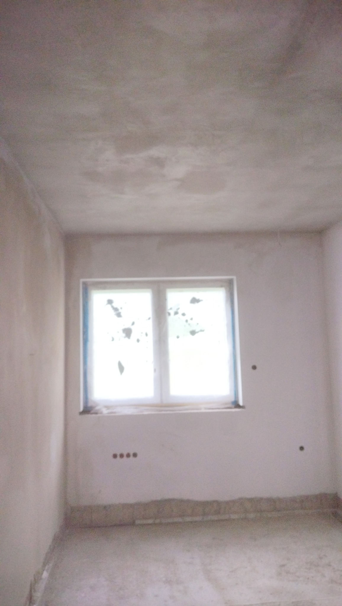 Surowe, jasne wnętrze pokoju po wstępnych pracach wykończeniowych, z oknem zabezpieczonym folią i ścianami pokrytymi szarym tynkiem, widoczne gniazdka elektryczne i nierówności przy podłodze.