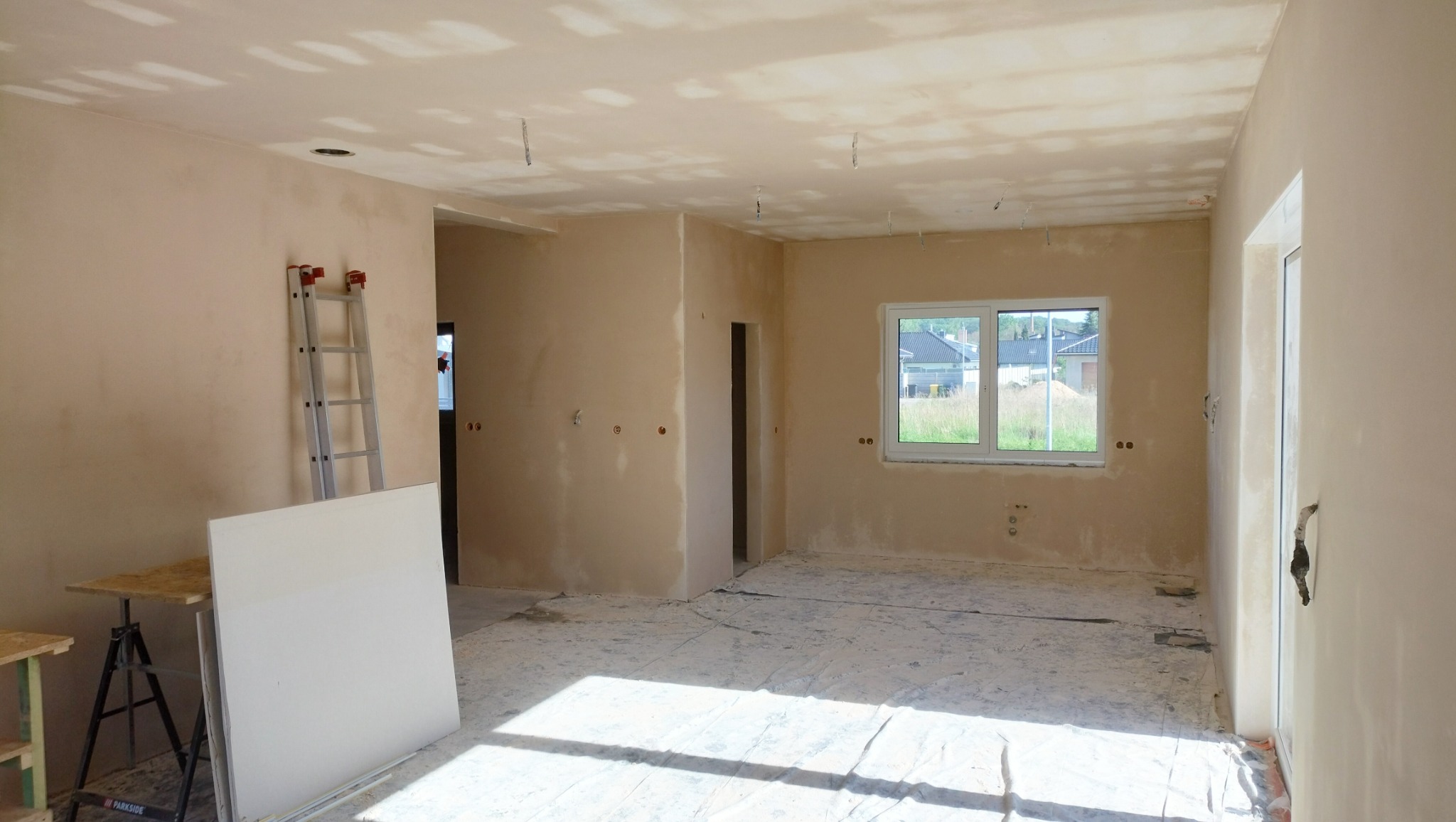 Pokój w trakcie remontu: ściany po wstępnym wyrównaniu, widoczne ślady po gładzi, zabezpieczona folią podłoga, drabina oparta o ścianę, płyta gipsowa na stole warsztatowym.