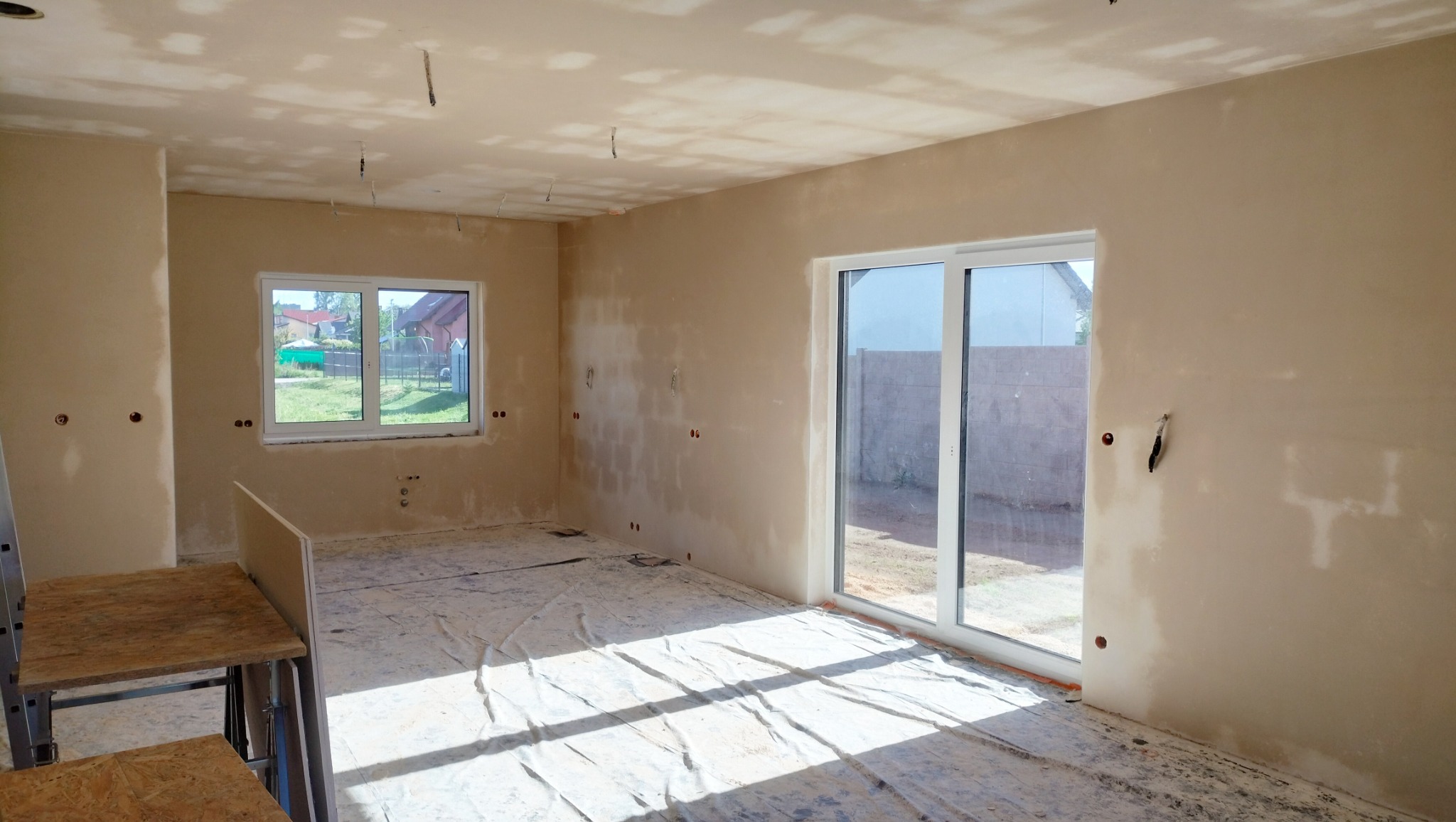 Pokój w stanie deweloperskim po wstępnym tynkowaniu maszynowym, z widocznymi śladami po gniazdkach elektrycznych, osłoniętą podłogą i jasnym światłem słonecznym wpadającym przez okno balkonowe.