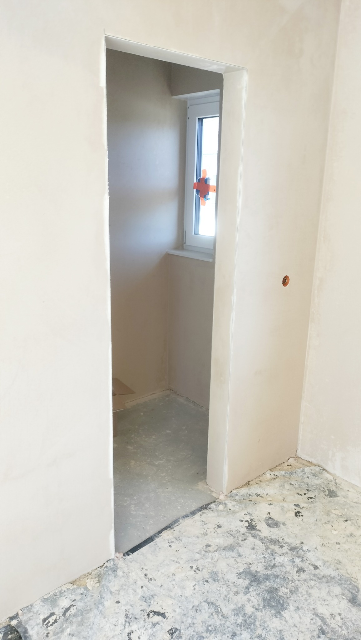Świeżo otynkowane wnętrze z widocznym oknem zabezpieczonym pomarańczowym krzyżakiem, surowa podłoga w nieukończonym pomieszczeniu.