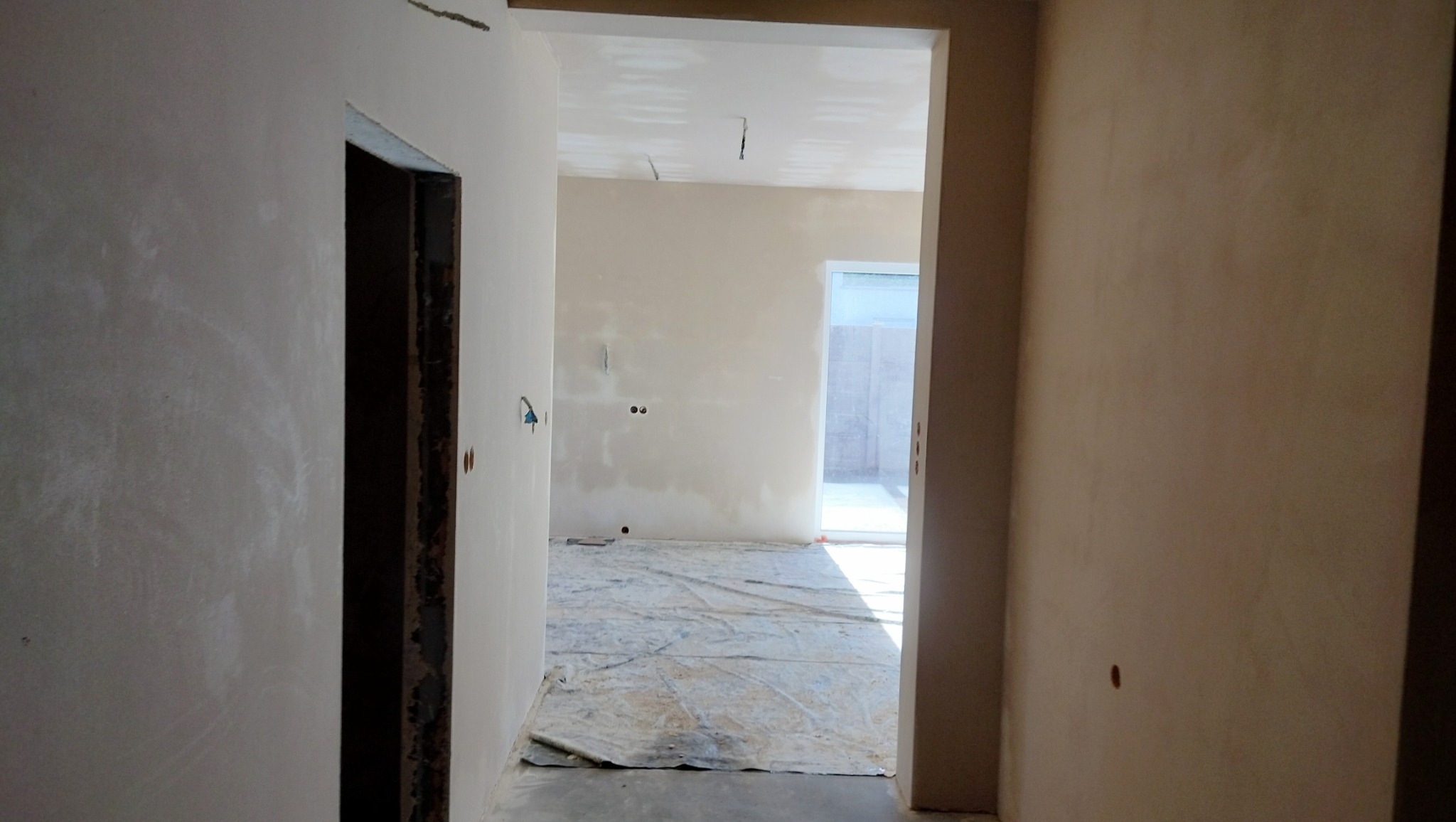 Świeżo otynkowane wnętrze w trakcie wykańczania, widok z korytarza na pomieszczenie z zabezpieczoną podłogą, ściany w jasnym kolorze przed malowaniem, widoczne puszki elektryczne.