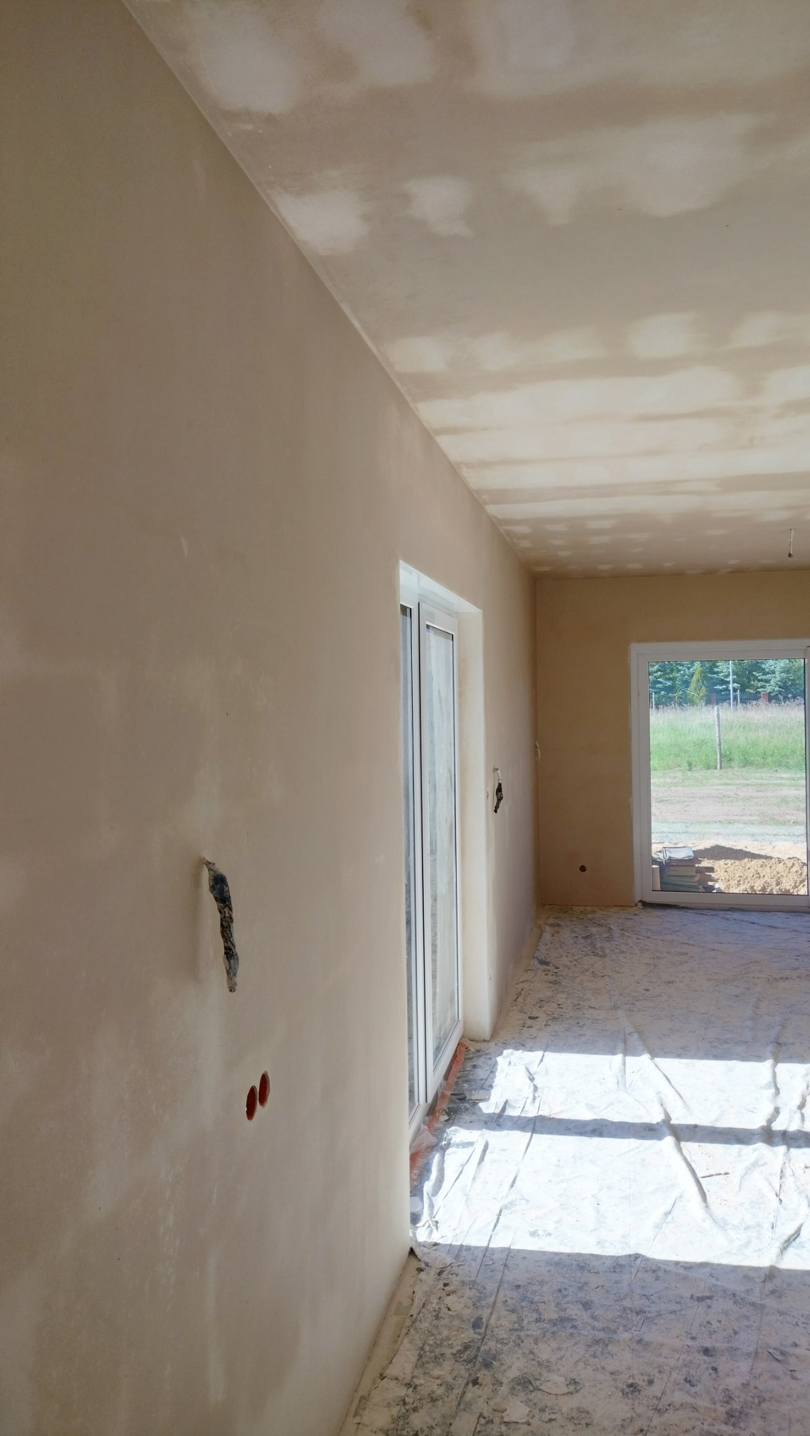 Wnętrze w stanie deweloperskim po wstępnym wyrównaniu ścian i sufitu, widoczne niedoskonałości powierzchni i zakryta folią podłoga, przygotowane otwory pod instalacje elektryczne.