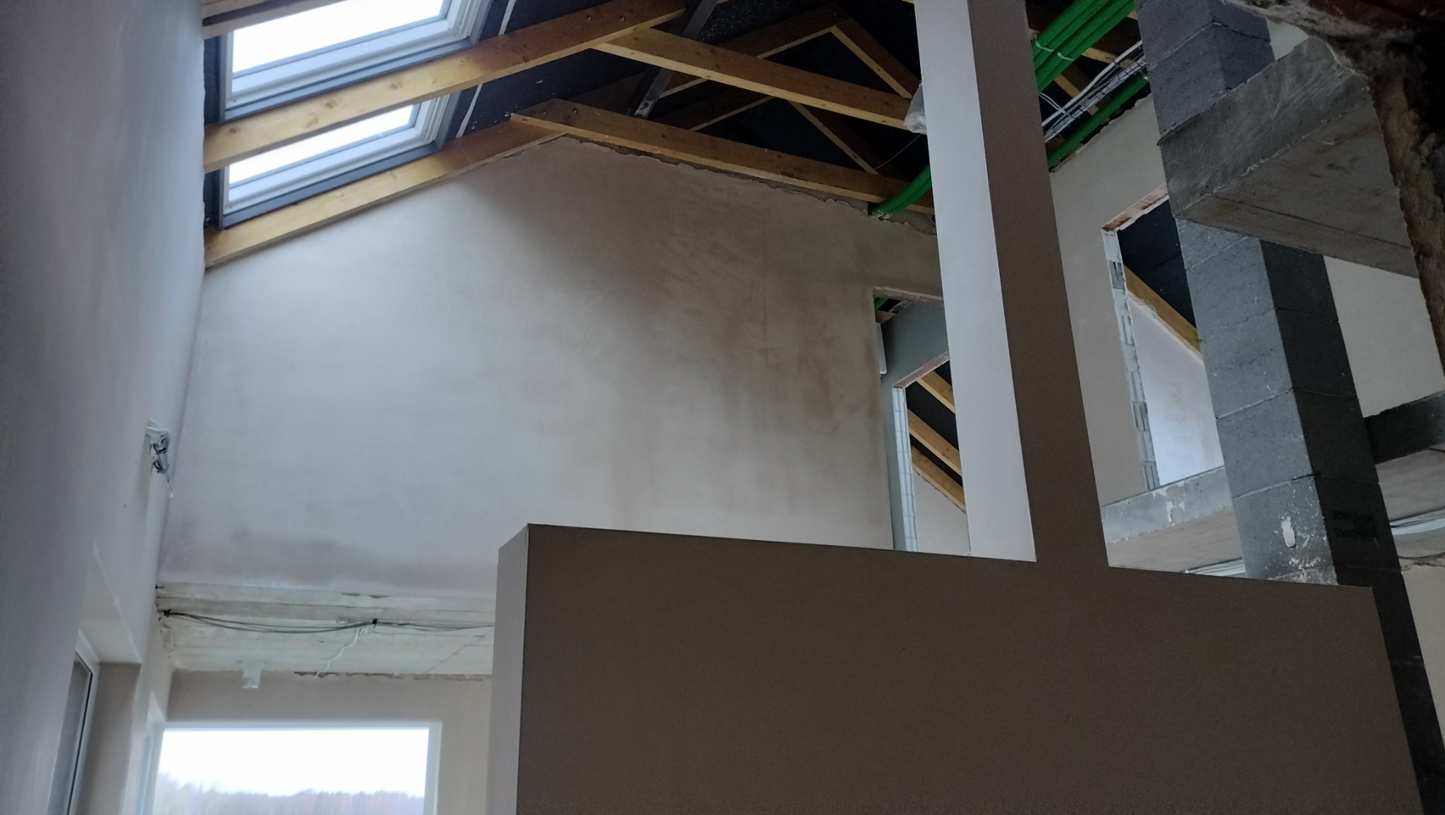 Wnętrze w trakcie prac wykończeniowych: ściany po nałożeniu gładzi, widoczne okna dachowe, drewniana konstrukcja dachu, instalacja elektryczna pod sufitem oraz fragmenty konstrukcji nośnej budynku.
