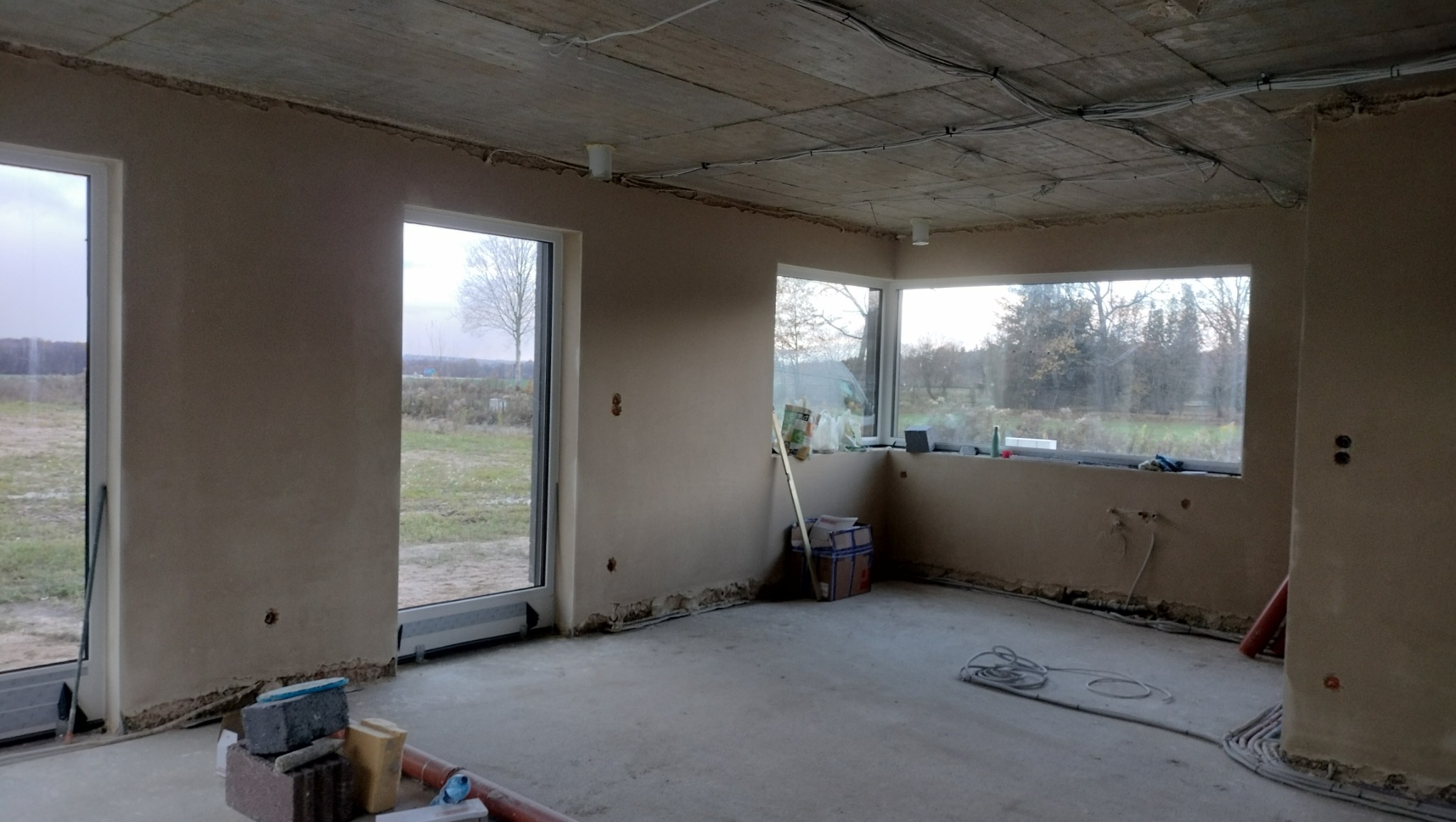 Surowe wnętrze domu w trakcie wykańczania: ściany pokryte świeżym tynkiem, widoczne przewody elektryczne na suficie, okna z widokiem na zewnątrz, materiały budowlane i narzędzia na podłodze.