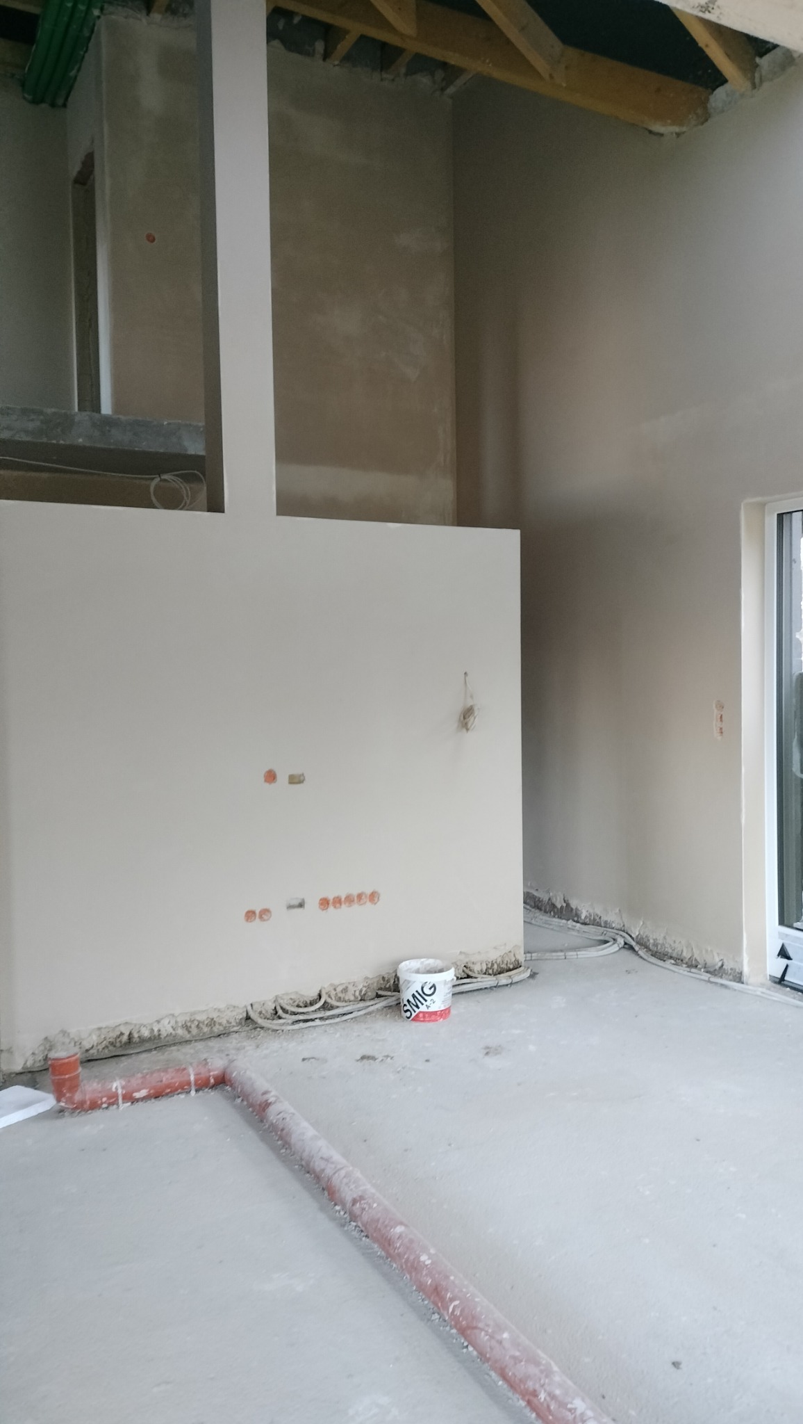 Wnętrze w trakcie prac wykończeniowych: ściany po nałożeniu gładzi, widoczne gniazdka elektryczne, rury w podłodze, wiadro z farbą SMIG przy ścianie. Surowy stan poddasza z widoczną konstrukcją dachu.