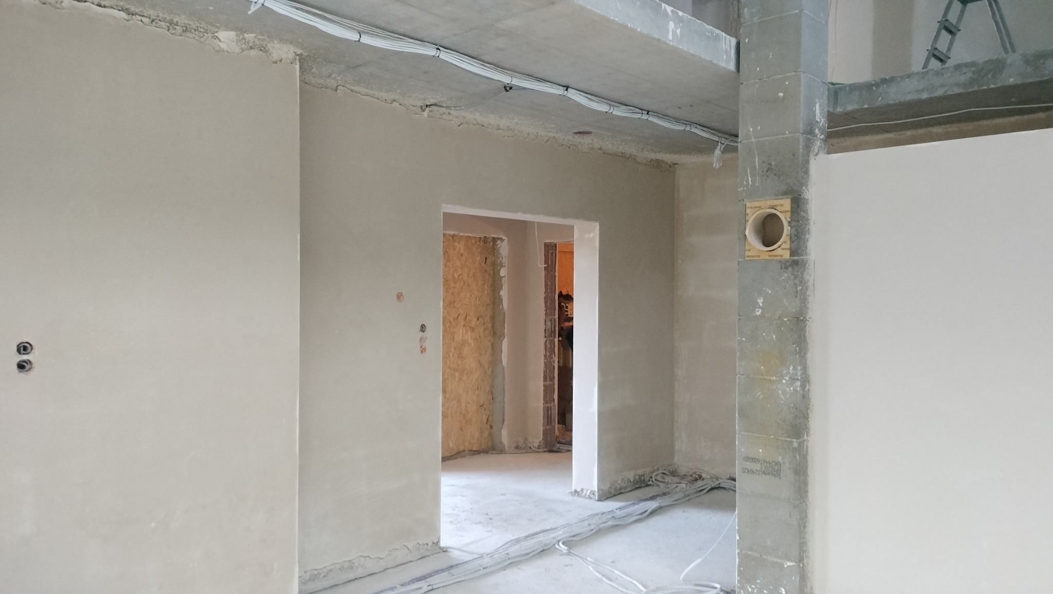 Wnętrze w trakcie prac wykończeniowych: ściany pokryte świeżym tynkiem maszynowym, widoczne przewody elektryczne pod sufitem i przy podłodze, surowy betonowy strop, otwór wentylacyjny w słupie...