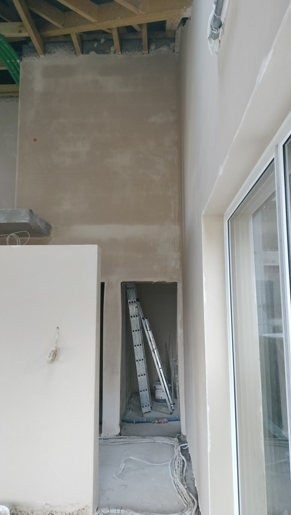 Wnętrze w trakcie prac wykończeniowych, ściany pokryte świeżym tynkiem, widoczna drabina w przejściu, instalacja elektryczna ułożona na podłodze, surowy stan poddasza.
