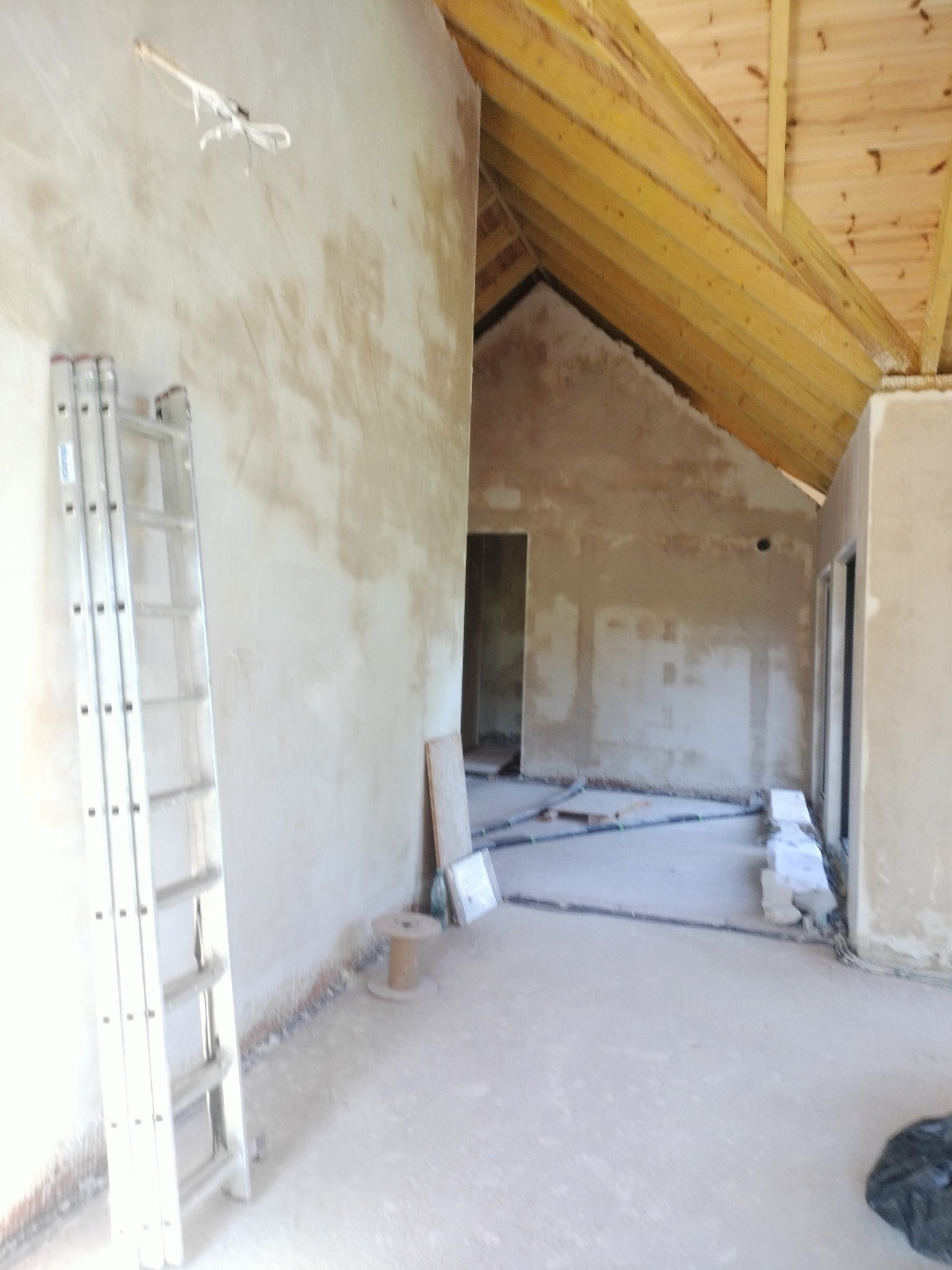 Wnętrze w trakcie prac wykończeniowych: ściany pokryte tynkiem, drewniany strop, drabina oparta o ścianę, instalacje na podłodze. Widoczny postęp prac.