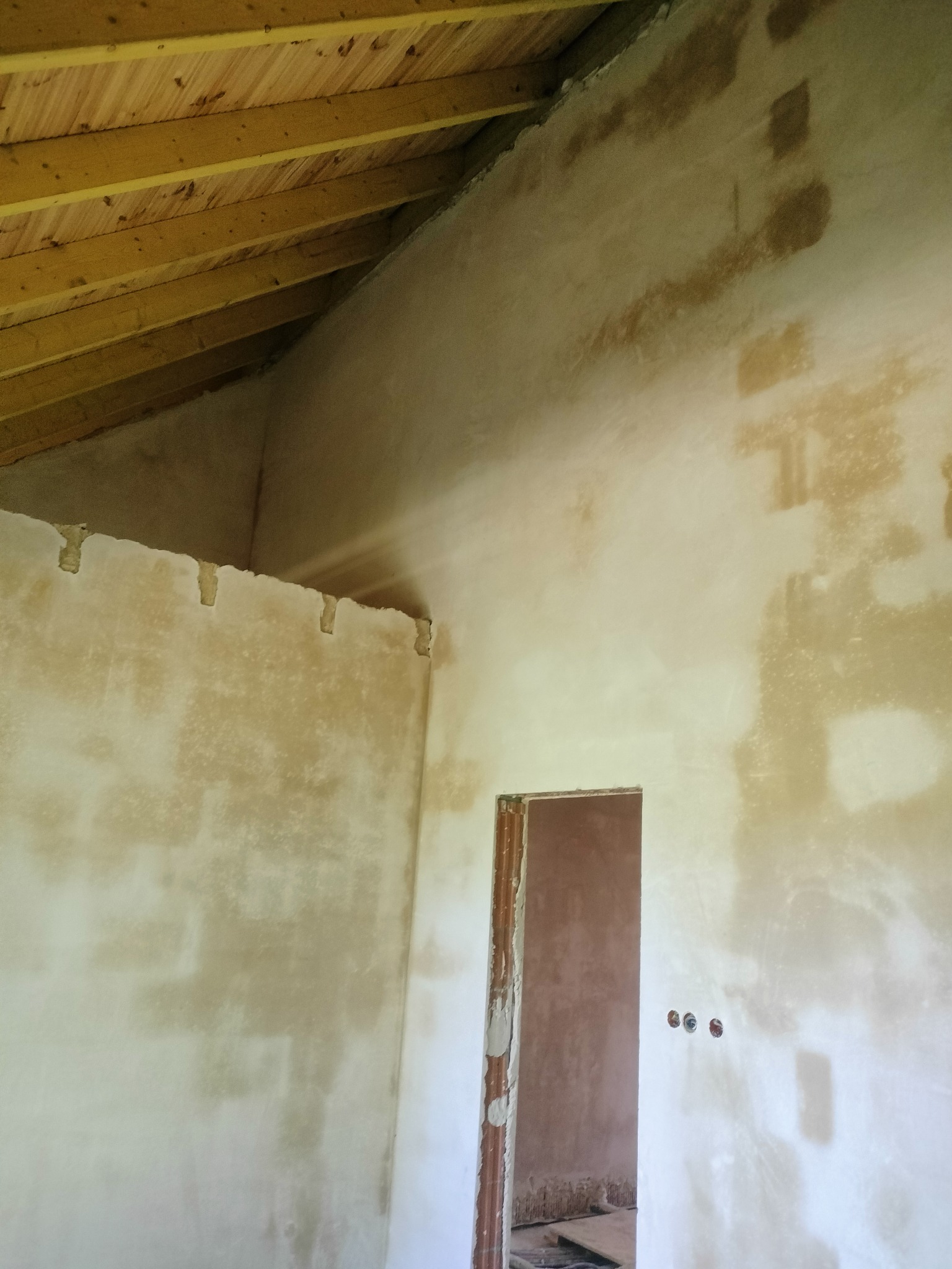 Wnętrze w trakcie prac wykończeniowych: surowy tynk na ścianach, drewniany strop, widoczne puszki elektryczne, przygotowany otwór drzwiowy. W tle widoczny fragment podłogi.