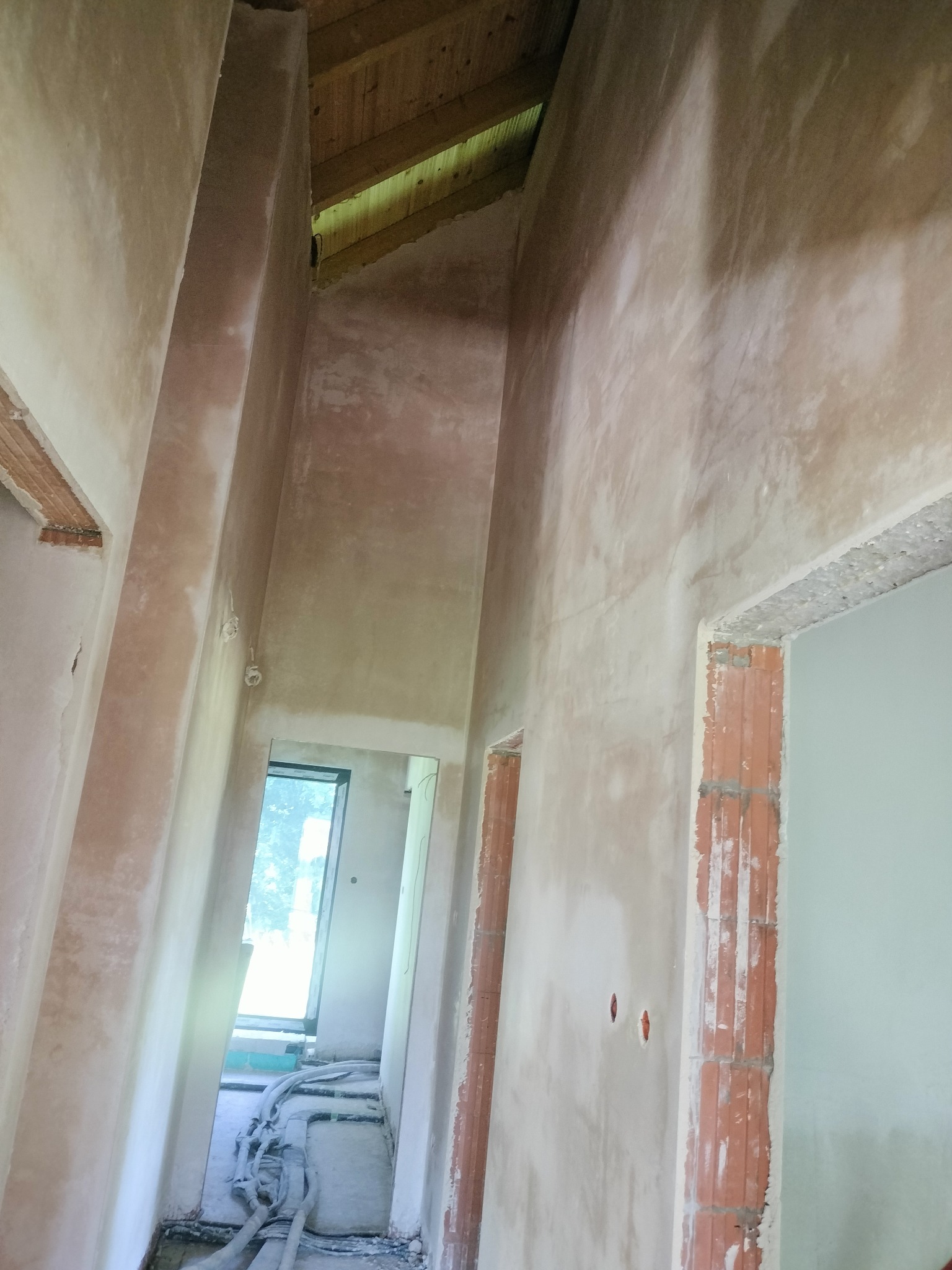 Wnętrze domu w trakcie prac wykończeniowych: surowe ściany pokryte tynkiem, widoczne instalacje na podłodze, drewniane elementy konstrukcyjne pod dachem.