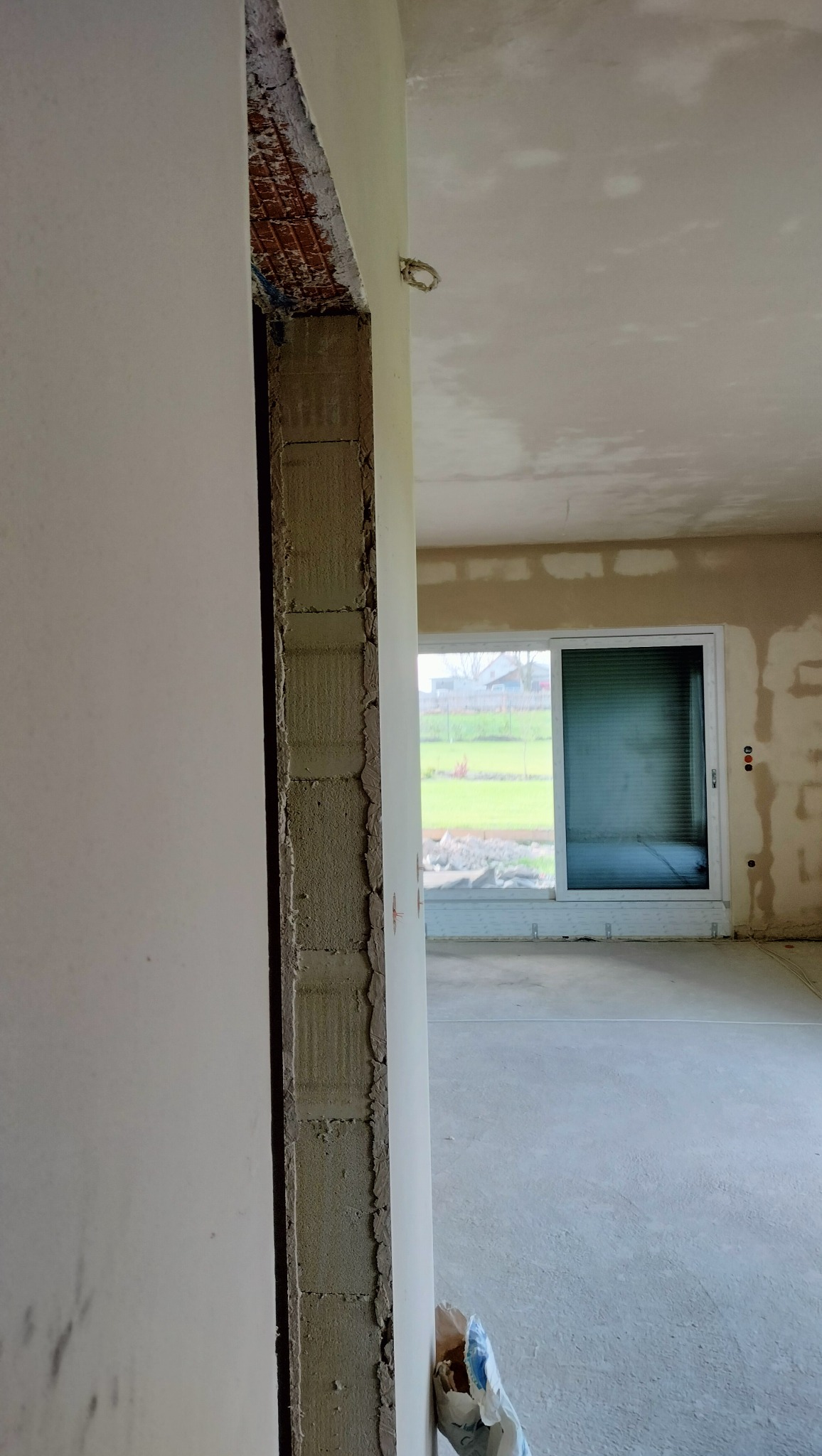 Surowy stan ścian wewnątrz remontowanego pomieszczenia. Widoczne ślady po tynkowaniu i przygotowaniu do dalszych prac wykończeniowych, okno przesuwne na wprost.