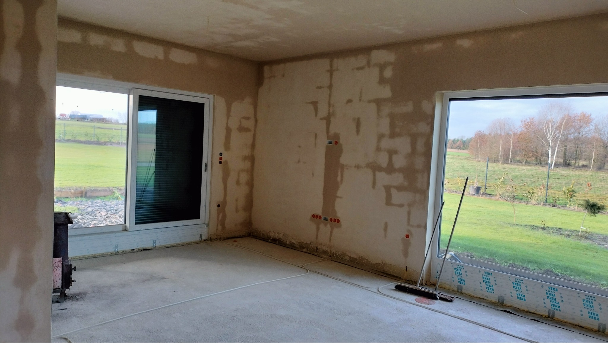 Wnętrze w trakcie tynkowania: ściany pokryte tynkiem, widoczne instalacje elektryczne, okna z widokiem na zewnątrz. Podłoga betonowa, przygotowana do dalszych prac wykończeniowych.