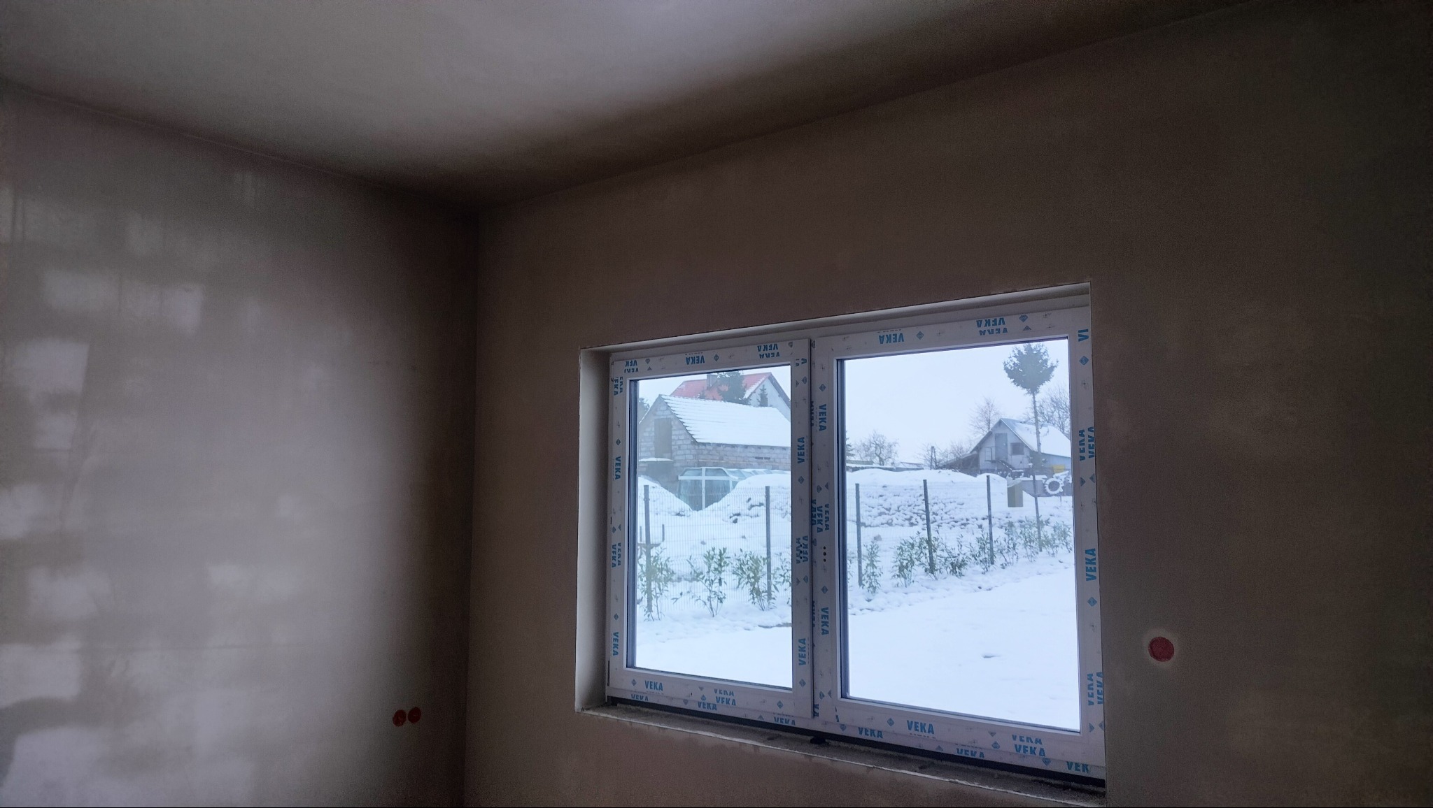 Pokój w trakcie prac wykończeniowych: świeżo położone gładzie na ścianach i suficie, białe okno z widokiem na zimowy krajobraz za oknem. Widoczne detale wykończenia.