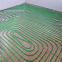 Zielone rury ogrzewania podłogowego ułożone na folii z czerwoną siatką, widoczne napisy 'IZOTAR' na folii, instalacja w trakcie realizacji.