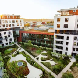 Widok z góry na wewnętrzny dziedziniec osiedla mieszkaniowego z nowoczesnym ogrodem, ścieżkami z jasnego żwiru i zielonymi nasadzeniami, w tle budynki mieszkalne.