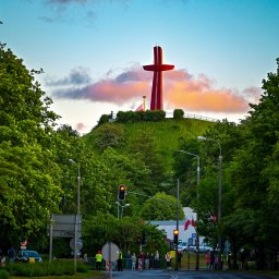 PBIKTZ Irbud sp z o.o. - Czerwony krzyż na zielonym wzgórzu, otoczony drzewami, widoczny z ulicy z sygnalizacją świetlną i przechodniami, pod błękitnym niebem z różowymi chmurami o zmierzchu.
