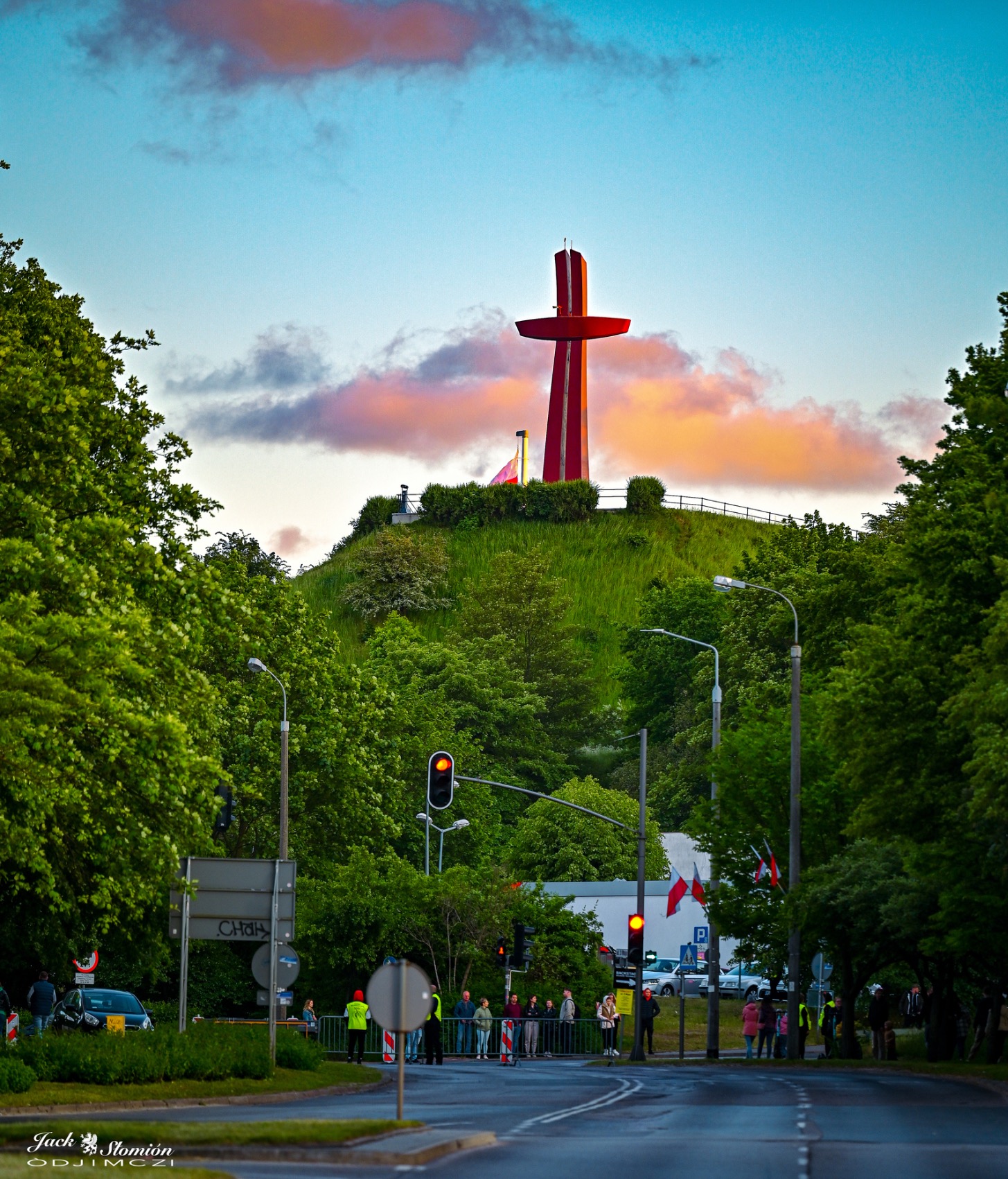 Czerwony krzyż na zielonym wzgórzu, otoczony drzewami, widoczny z ulicy z sygnalizacją świetlną i przechodniami, pod błękitnym niebem z różowymi chmurami o zmierzchu.