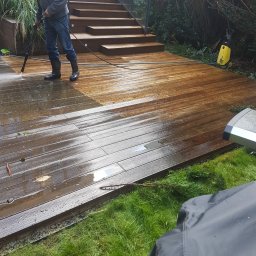 Ogrod Marzen - Mężczyzna w gumowych butach czyści drewniany taras myjką ciśnieniową, widoczna różnica w kolorze między umytą a brudną częścią, schody z drewnianymi stopniami w tle.