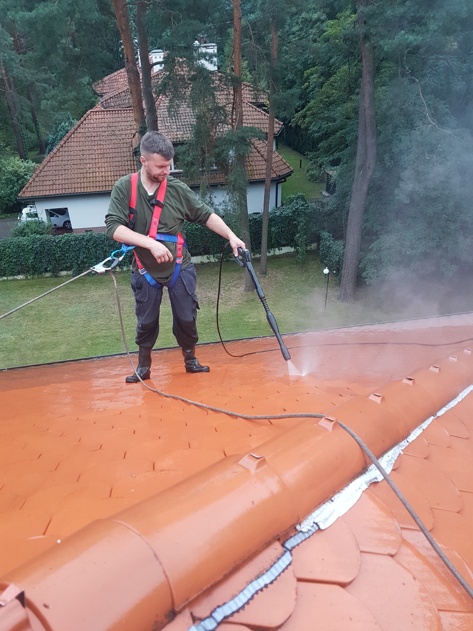 Pracownik w uprzęży zabezpieczającej czyści dach z ceramicznej dachówki przy użyciu myjki ciśnieniowej, w tle widoczny dom i zieleń.