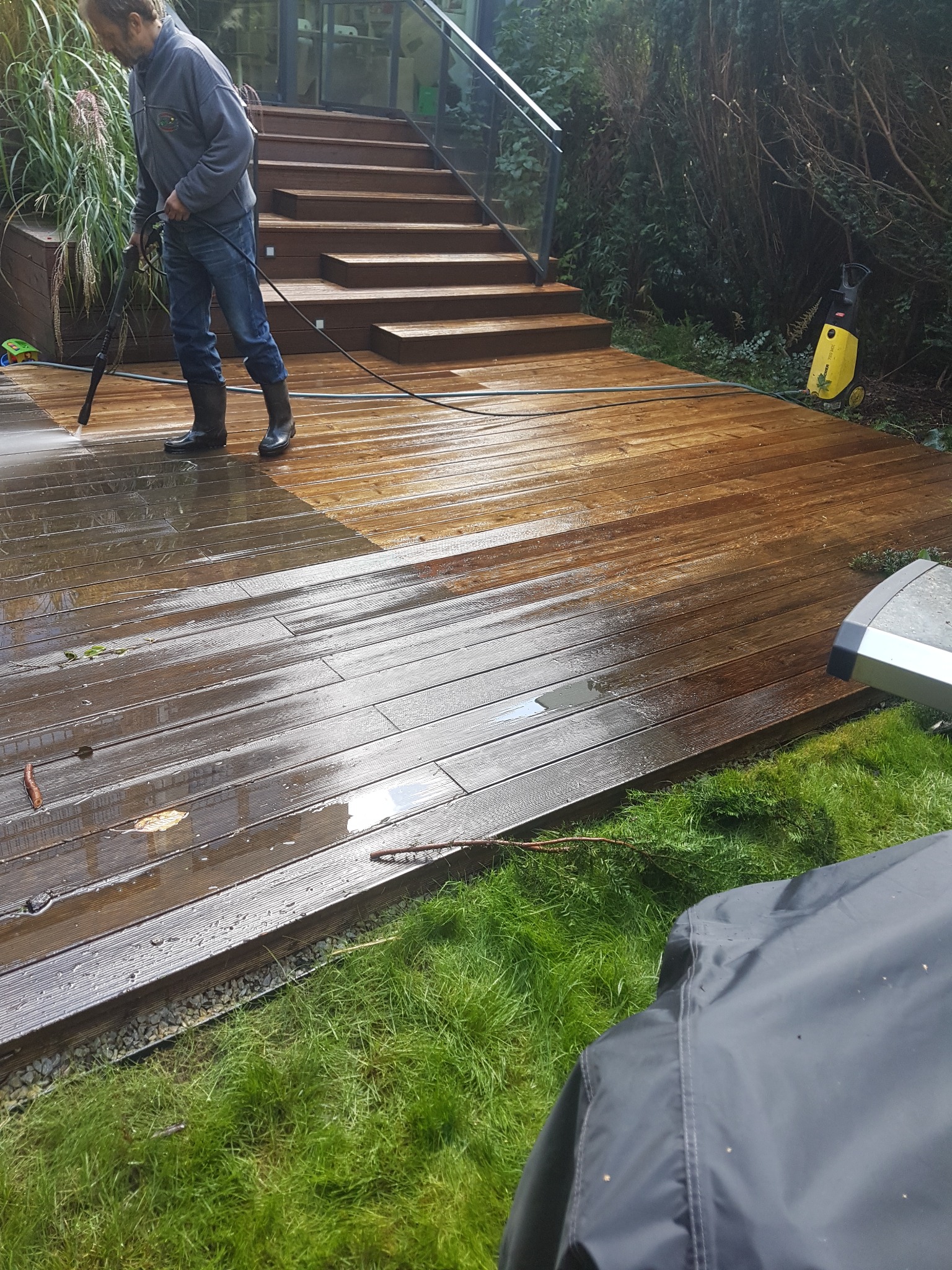 Mężczyzna w gumowych butach czyści drewniany taras myjką ciśnieniową, widoczna różnica w kolorze między umytą a brudną częścią, schody z drewnianymi stopniami w tle.