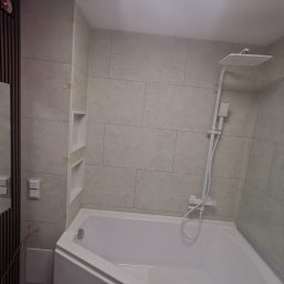 ANDRZEJ DOBRZE CI ZROBI USŁUGI REMONTOWE - Nowoczesna, narożna wanna w jasnej łazience z beżowymi płytkami, minimalistyczną armaturą i wnękami w ścianie, oświetlona światłem z okna dachowego.