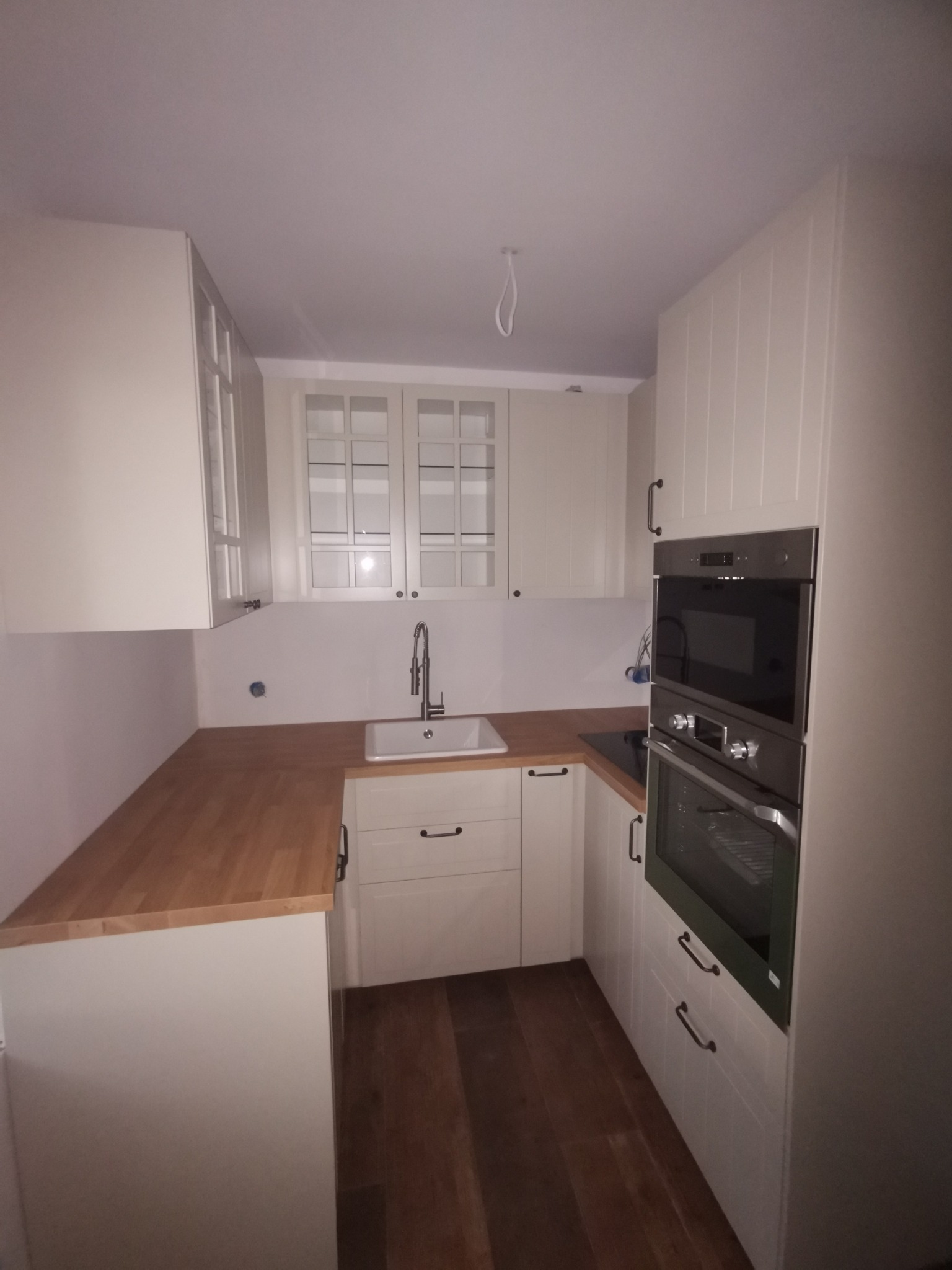 Jasna kuchnia w stylu skandynawskim z drewnianym blatem i podłogą, widoczne zabudowane piekarniki i szafki z przeszklonymi frontami.