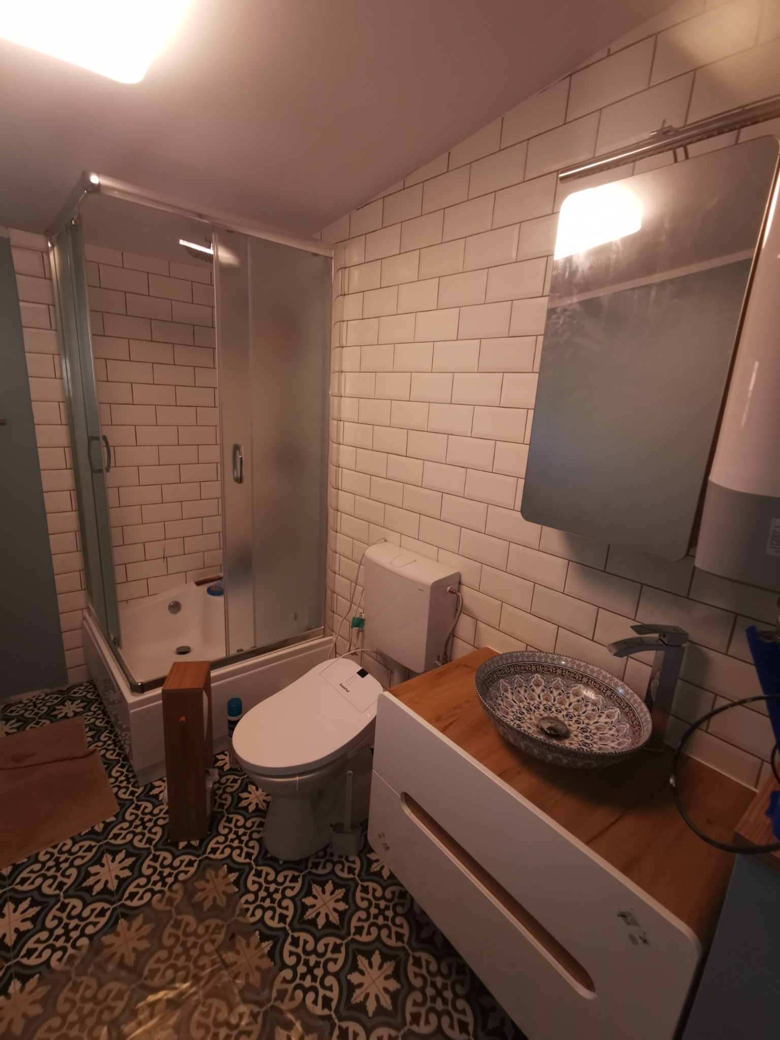 Ukończona łazienka z kabiną prysznicową, toaletą myjącą, umywalką nablatową w orientalne wzory i białą szafką pod umywalką, wykończona białymi płytkami imitującymi cegłę i wzorzystą podłogą.