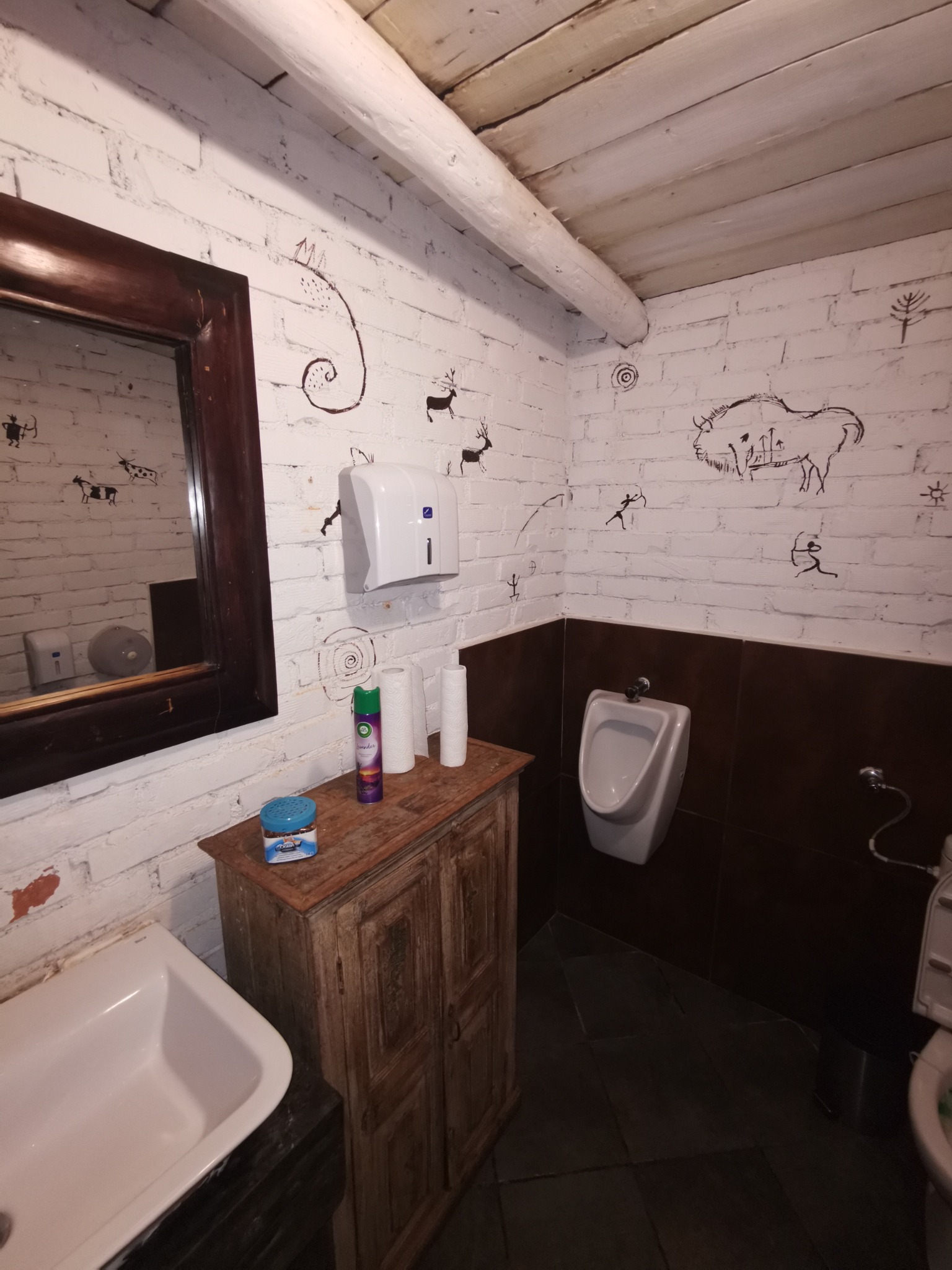 Nietypowa łazienka z białymi cegłami ozdobionymi rysunkami naskalnymi zwierząt, drewnianą szafką pod umywalką i lustrem w ciemnej ramie.