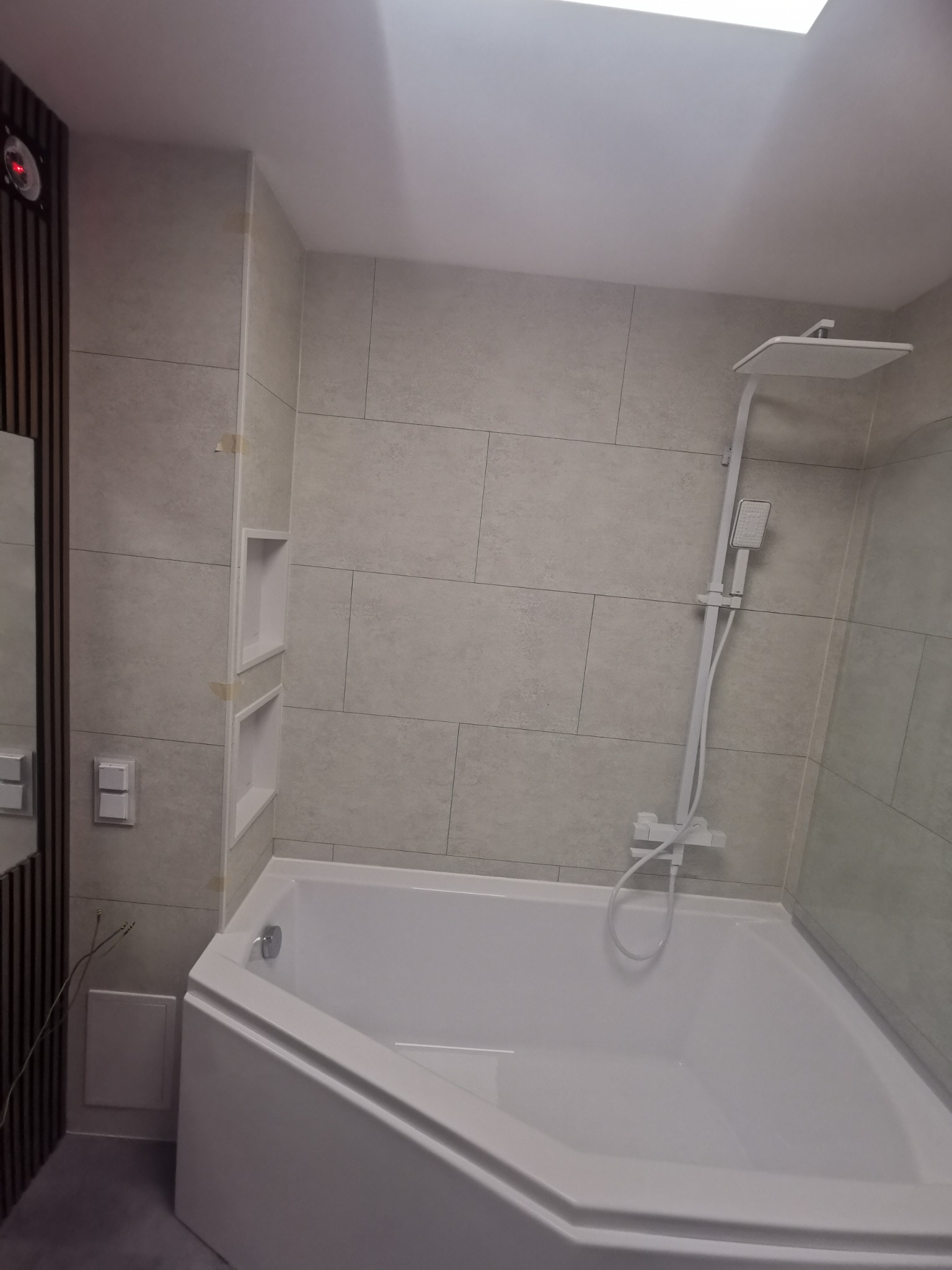 Nowoczesna, narożna wanna w jasnej łazience z beżowymi płytkami, minimalistyczną armaturą i wnękami w ścianie, oświetlona światłem z okna dachowego.