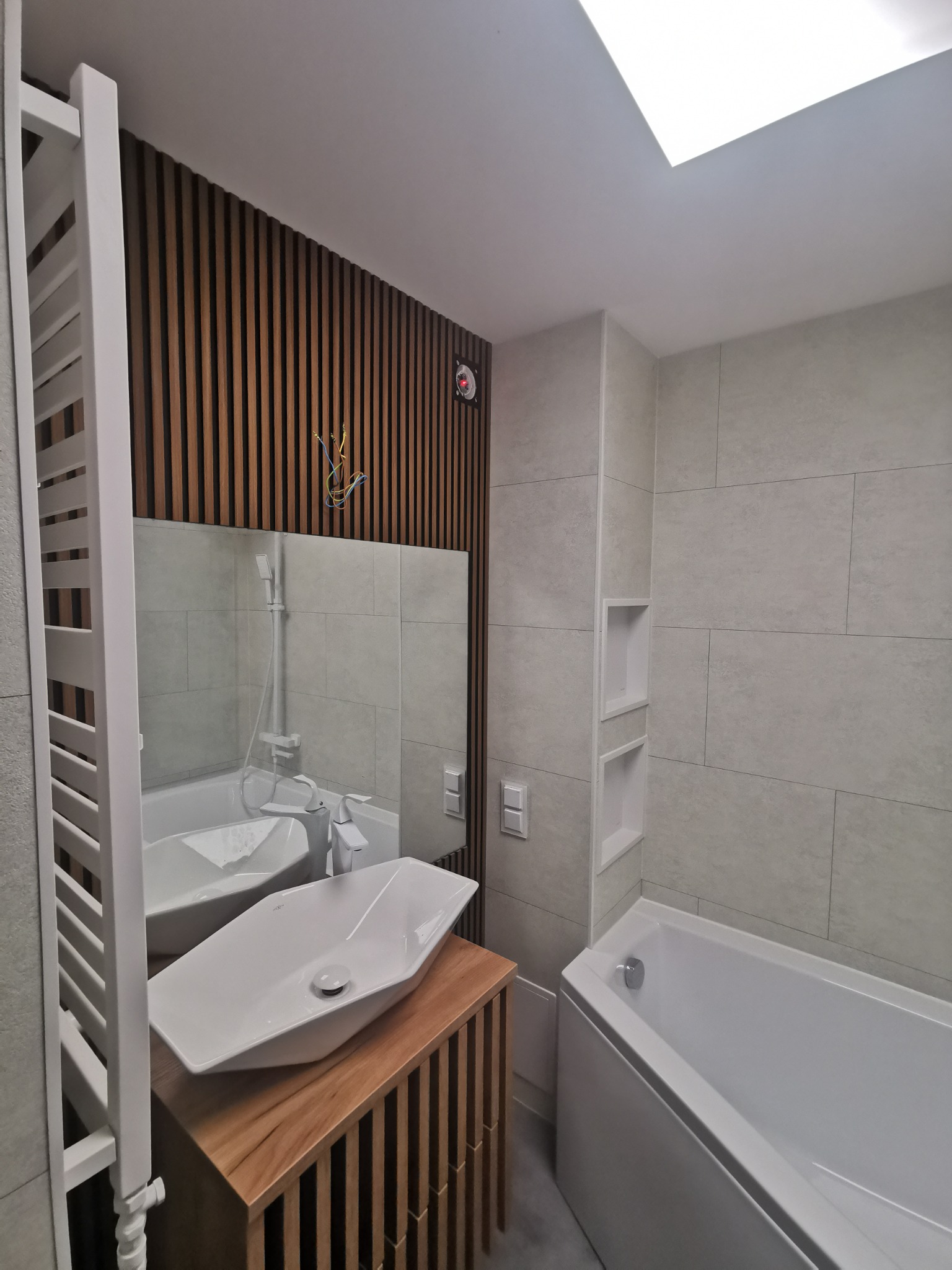 Nowoczesna łazienka z drewnianą ścianą z pionowych listew, asymetryczną umywalką nablatową i grzejnikiem drabinkowym w kolorze białym.