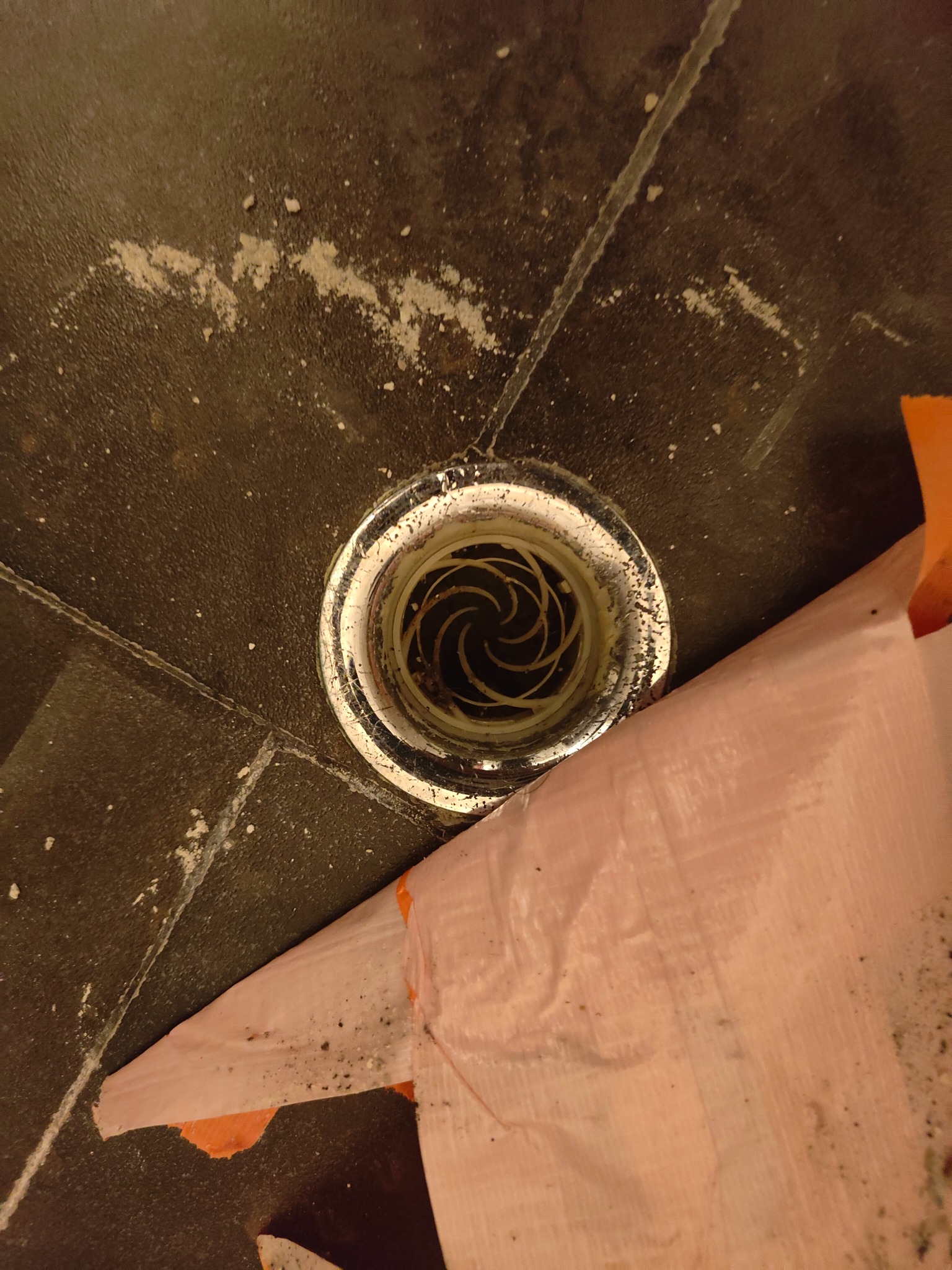 Ujęcie z góry na odpływ podłogowy w ciemnych płytkach, częściowo zakryty folią ochronną podczas prac wykończeniowych, widoczne resztki zaprawy.