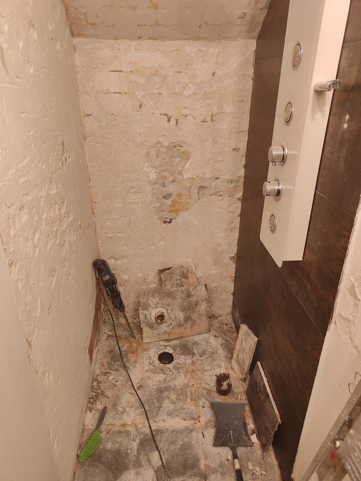 Demontaż starej armatury w trakcie remontu łazienki, widoczna ściana z resztkami starej glazury, panel prysznicowy i narzędzia: młot udarowy oraz szpachelka.