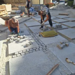 Dwóch mężczyzn w trakcie układania dużych płyt kamiennych z obrzeżami z kostki na placu budowy, widoczne narzędzia i materiały budowlane w tle.