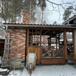 Drewniana altana z ceglanym kominem w zimowej scenerii, na pierwszym planie taczka i drabina oparta o ścianę, śnieg na ziemi.
