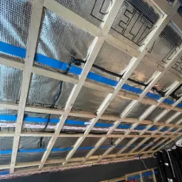 Konstrukcja drewniana dachu z izolacją termiczną i folią paroizolacyjną z nadrukiem, zabezpieczona niebieską taśmą, widoczne przewody elektryczne.