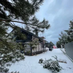 Niedokończony dom szkieletowy w zimowej scenerii; widoczne rusztowania, folia izolacyjna i ślady na śniegu, z gałęziami sosny w pierwszym planie.