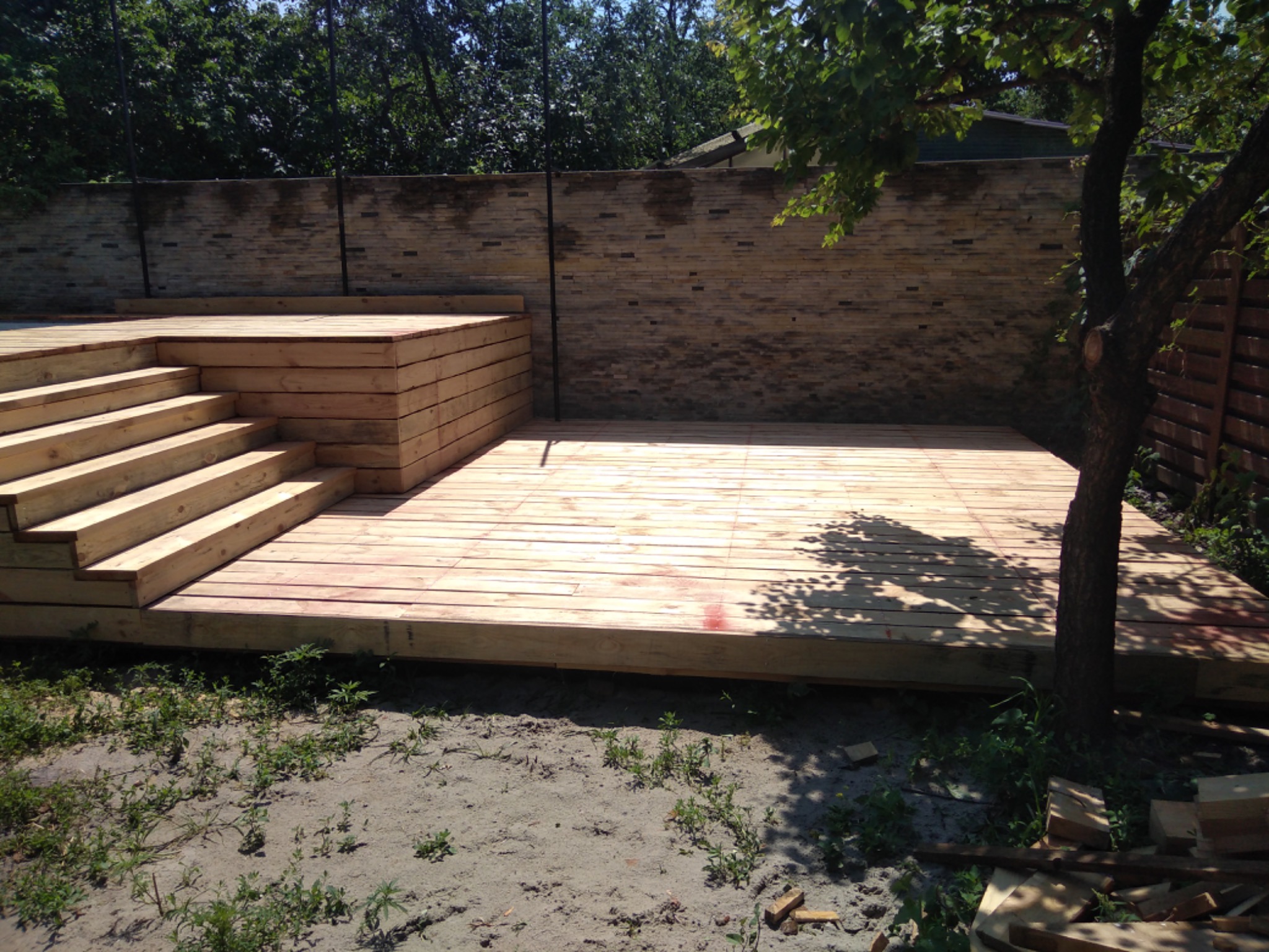 Nowo wybudowany taras drewniany z schodami, surowe drewno, widoczne ślady po cięciu, na tle ceglanego muru i drzewa rzucającego cień.