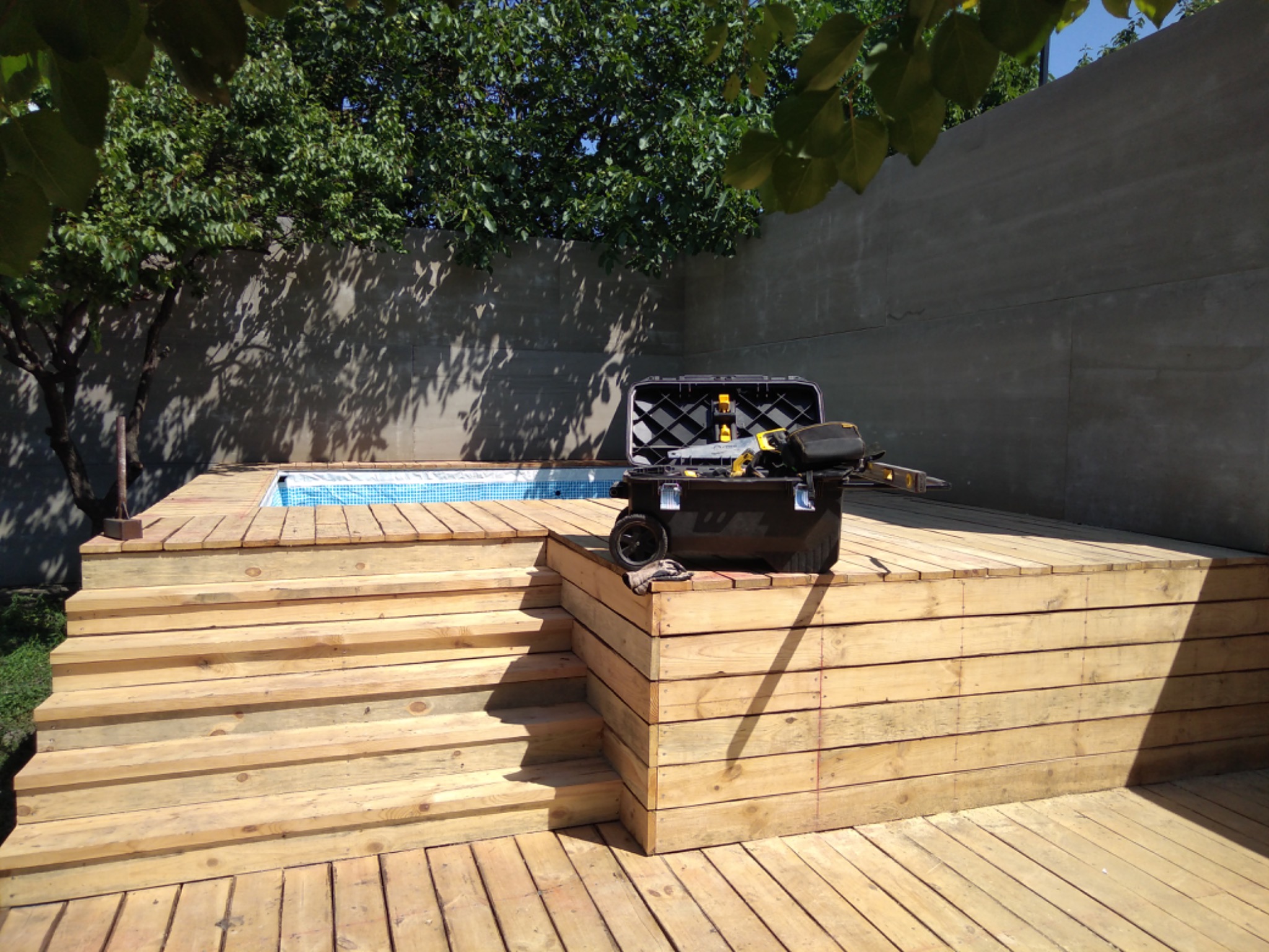 Nowy taras z jasnego drewna z widocznymi schodami i basenem w tle, obok skrzynka narzędziowa.