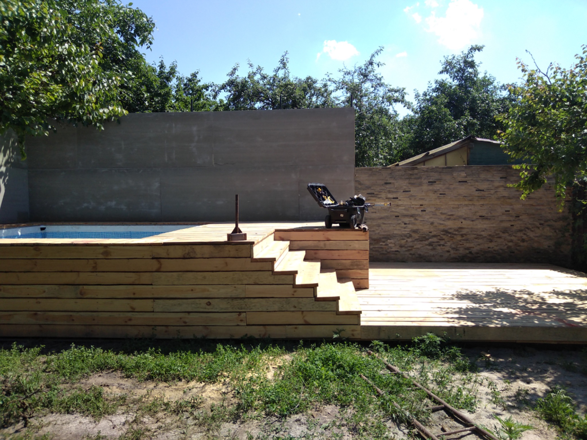 Nowo wybudowany taras z jasnego drewna z niskimi schodami, przylegający do basenu i otoczony zielenią, z widoczną skrzynką narzędziową na górze.