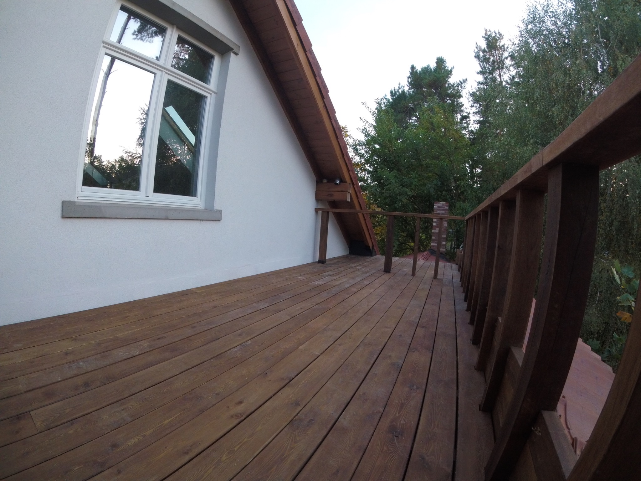 Przestronny taras z brązowych desek, przylegający do białej ściany domu z oknem, z drewnianą balustradą i widokiem na drzewa w tle.