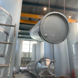 Montaż dużych, cylindrycznych zbiorników ze stali nierdzewnej w hali produkcyjnej, jeden zbiornik podnoszony za pomocą dźwigu, widoczne spawarki i inne zbiorniki w tle.