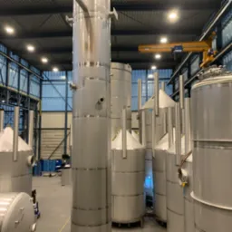 Wnętrze hali produkcyjnej z szeregiem dużych, pionowych zbiorników ze stali nierdzewnej, niektóre z stożkowymi górnymi częściami, oświetlonych przez lampy sufitowe i z żółtym dźwigiem widocznym w tle.