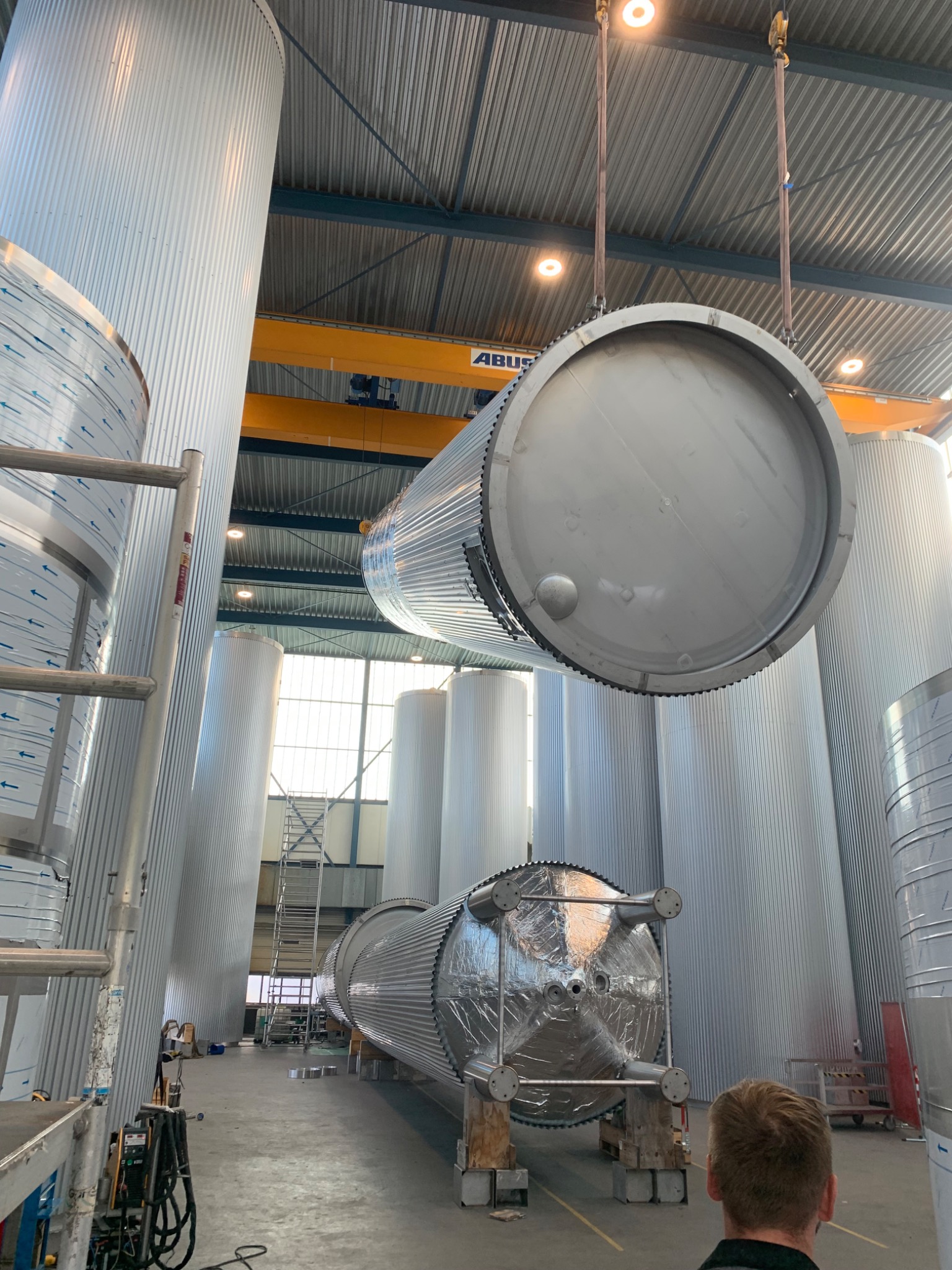 Montaż dużych, cylindrycznych zbiorników ze stali nierdzewnej w hali produkcyjnej, jeden zbiornik podnoszony za pomocą dźwigu, widoczne spawarki i inne zbiorniki w tle.