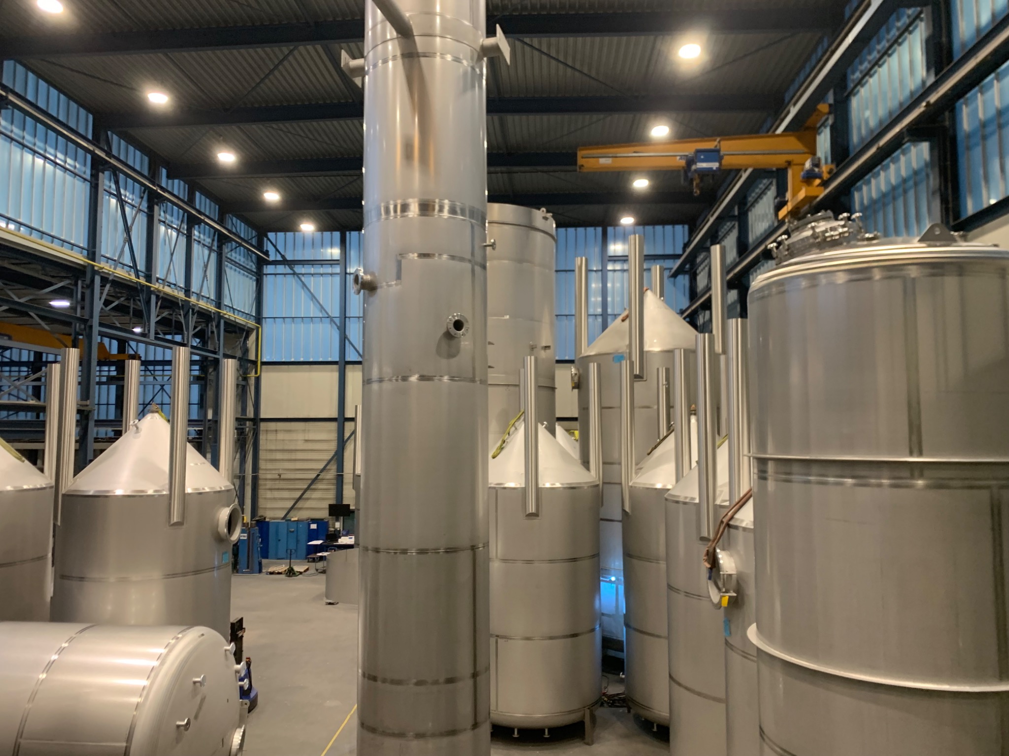 Wnętrze hali produkcyjnej z szeregiem dużych, pionowych zbiorników ze stali nierdzewnej, niektóre z stożkowymi górnymi częściami, oświetlonych przez lampy sufitowe i z żółtym dźwigiem widocznym w tle.