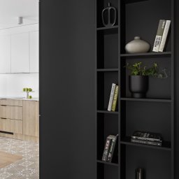 Modul-AR Architektura Eliza Poręba - Częściowy widok nowoczesnego mieszkania: czarny regał z dekoracjami (wazony, książki, roślina), fragment kuchni z drewnianymi frontami i blatem, podłoga z płytek wzorzystych i parkietu.