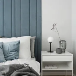 Sypialnia z niebieskim, tapicerowanym panelem zagłówkowym, białą pościelą, szarą narzutą w kratę i białą szafką nocną z lampką i dekoracjami.