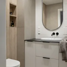 Nowoczesna łazienka z owalnym lustrem, białą umywalką nablatową z czarną armaturą i szafką pod umywalkową z szufladami, obok toaleta podwieszana i drewniana szafka z półkami.