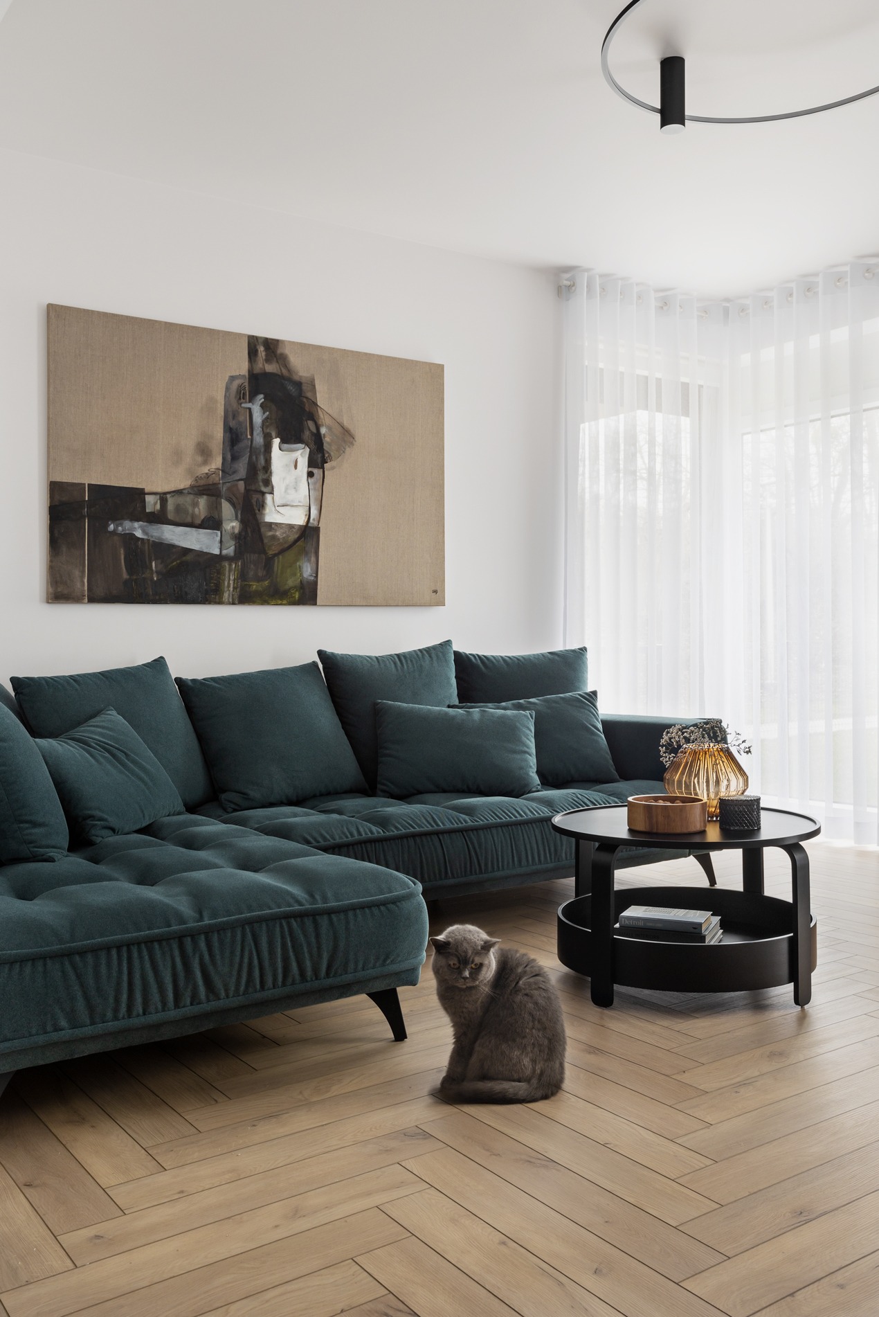 Salon z ciemnozieloną sofą narożną, abstrakcyjnym obrazem na ścianie i szarym kotem siedzącym na parkiecie w jodełkę.